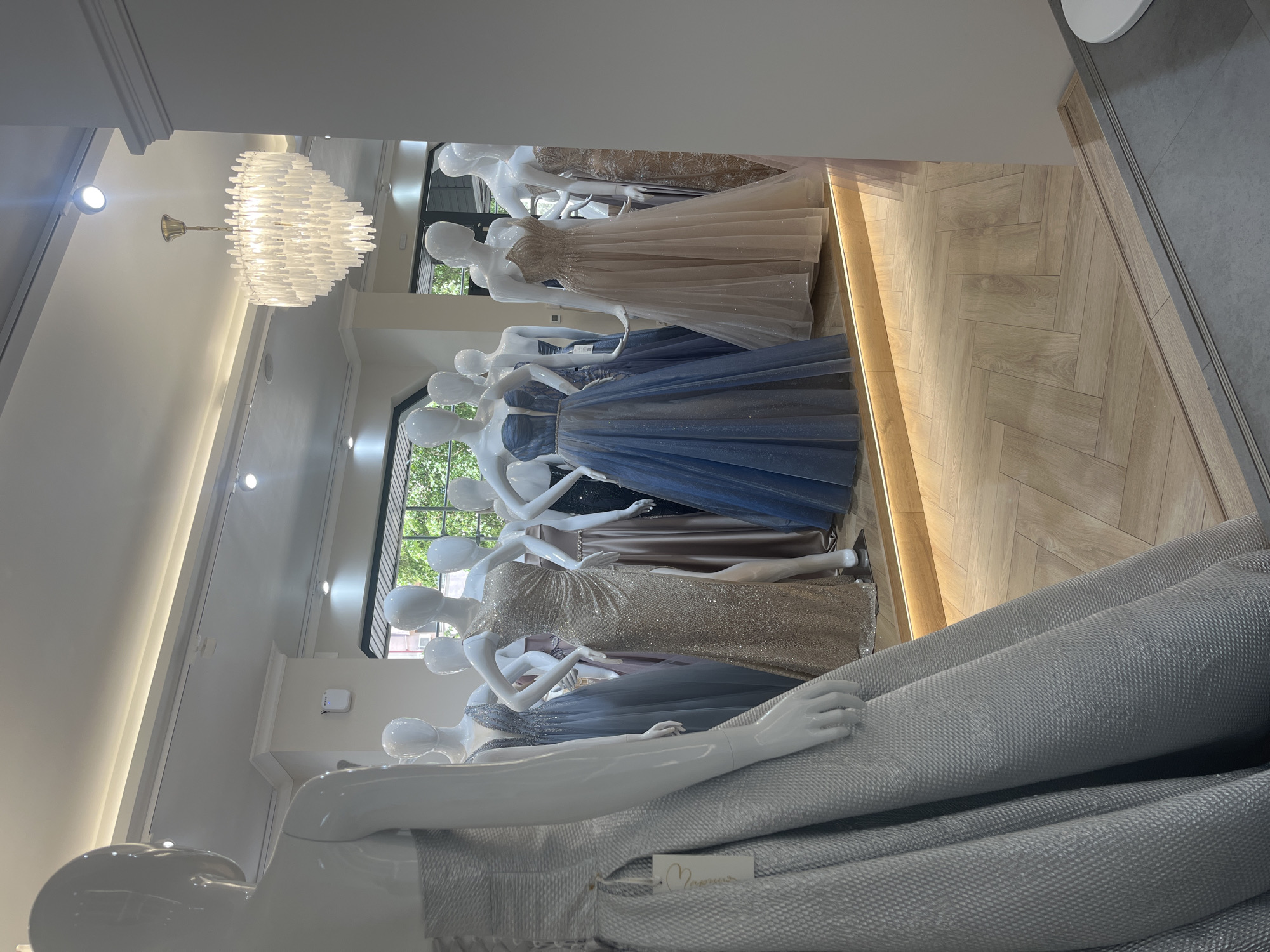 A retail setting showcasing formal gowns on mannequins. The location is Blagoevgrad, Bulgaria. The mannequins, all headless and white, are arranged in a row, each wearing a floor-length dress in various colors and designs including blue, gold, and silver. The gowns appear to be constructed from shimmering or textured fabrics. The floor is light-colored wood, possibly a herringbone pattern. A modern chandelier hangs from the ceiling. Natural light enters the space, filtering through windows behind the mannequins. One mannequin in the foreground is draped with a textured, light-colored fabric. A price tag or label is visible on the fabric.