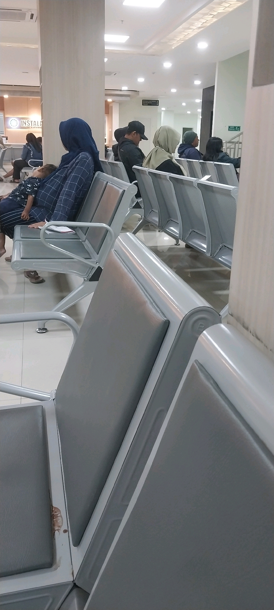 The image depicts a waiting area, likely a hospital or clinic, in Kebonagung, Indonesia. Rows of gray, metallic chairs with padded seats and backs are arranged facing a wall. Several individuals are seated, some with their heads bowed or resting. In the foreground, a person wearing a blue headscarf is holding a sleeping child. To the right, other individuals are seated, facing forward or to the side. The environment is indoors, with bright, artificial lighting from ceiling panels. A sign indicating "Jalan Keluar" (Exit) and an arrow pointing to the right is visible in the background, suggesting a public facility. The overall impression is one of a calm, routine scene of people waiting.
