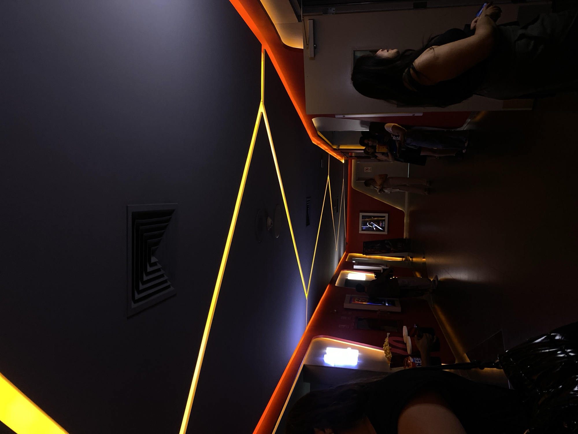 Here is a detailed description of the image:
**Scene:** The image shows the interior of what appears to be a modern cinema or entertainment venue, likely in an unknown city. The architectural design features sharp angles and neon lighting, giving it a futuristic look.
**People:** Several people are visible, including:
* One woman holding a phone.
* A group of people lined up, possibly waiting in a queue.
**Objects:** Key objects and architectural elements include:
* Neon lights in yellow and orange, outlining the walls and corridors.
* Modern ceiling vents.
* A wall mounted fan
* A display area with various items, possibly for refreshments or merchandise.
* A framed picture on the wall.
* Various displays on the wall
**Actions and Interactions:**
* Some individuals are standing or walking, suggesting movement within the venue.
* The woman is occupied with her phone, potentially communicating or using an app.
**Environment:**
* The lighting is dim overall, with the neon lights creating contrasting highlights and shadows.
* The space seems well-maintained and designed for a specific entertainment purpose.
The image lacks specific indicators of the city or precise location. However, the architectural details and lighting suggest a modern entertainment setting.