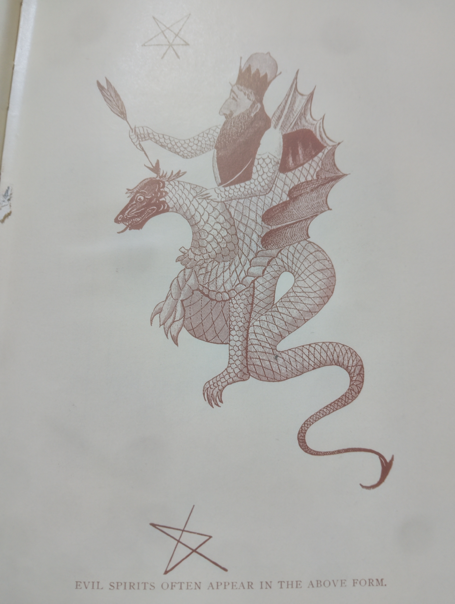The image shows an illustration in reddish-brown ink on a cream-colored background. At the top of the illustration is a five-pointed star with a circle around it. Below this, a stylized figure wearing a crown rides on the back of a winged, scaly dragon. The figure has a beard and holds a feather-like object in its right hand. The dragon has sharp claws, a long tail, and its mouth is open with its tongue extended. At the bottom of the illustration, there is another five-pointed star, and below that, the text "EVIL SPIRITS OFTEN APPEAR IN THE ABOVE FORM." The image depicts a mythological or symbolic scene rather than a real-world location. There is no information in the image that suggests a location in Alagbado, Nigeria.