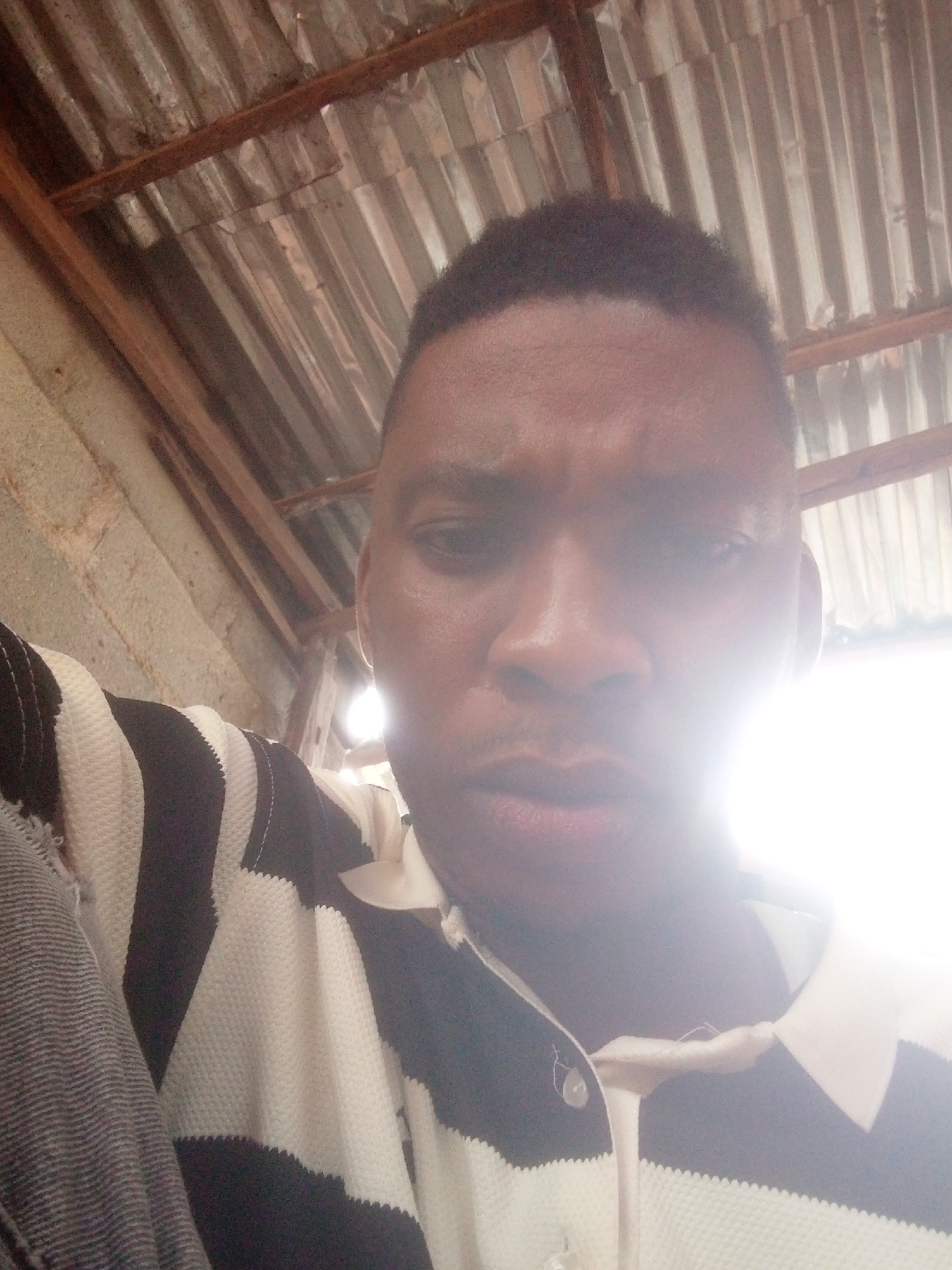 A man is indoors with a corrugated metal roof above him. He is wearing a black and white striped shirt with a white collar and is looking at the camera with a slightly furrowed brow. The light is bright, possibly from an opening in the roof. The location is Unknown, Unknown.