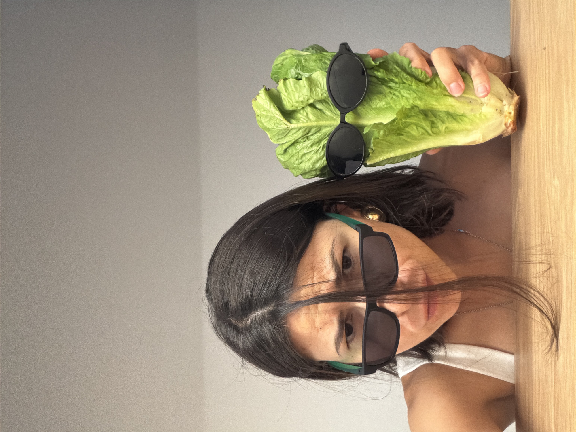 A woman with dark hair and sunglasses stands in front of a light gray wall and a light-colored wooden surface. She holds a head of romaine lettuce, also adorned with sunglasses, above her head. The location is not discernible from the image.