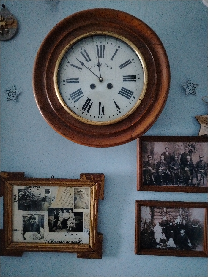A light blue, textured wall in Lezhnevo, Russia, displays a collection of framed items. Dominating the upper center is a large, circular wall clock with a dark wooden frame and a brass bezel. Its white face features black Roman numerals and the inscription "Le roy a Paris," with hands indicating approximately 10:10.
To the upper left of the clock, a small wooden cutout depicting stylized deer is visible, along with a white star-shaped decorative cutout. Another similar star is positioned to the right of the clock, and a third, partially visible star-shaped object is at the top right corner.
Below the clock, on the left side, a large, ornate rectangular wooden frame contains a collage of vintage black and white photographs and paper fragments. Visible elements within this collage include a photo of a young child with flowers, a portrait of a woman, a group of seated individuals, another woman, and text fragments with "ЯНВАРЬ" (January) and "1905."
To the right of the collage frame, two smaller, rectangular wooden frames are stacked vertically. The upper frame holds a sepia-toned group photograph of approximately ten men, some seated, some standing, dressed in what appears to be formal or military attire. The lower frame contains another sepia-toned group photograph depicting about ten to twelve individuals, including men, women, and children, posed outdoors or against a rustic backdrop.