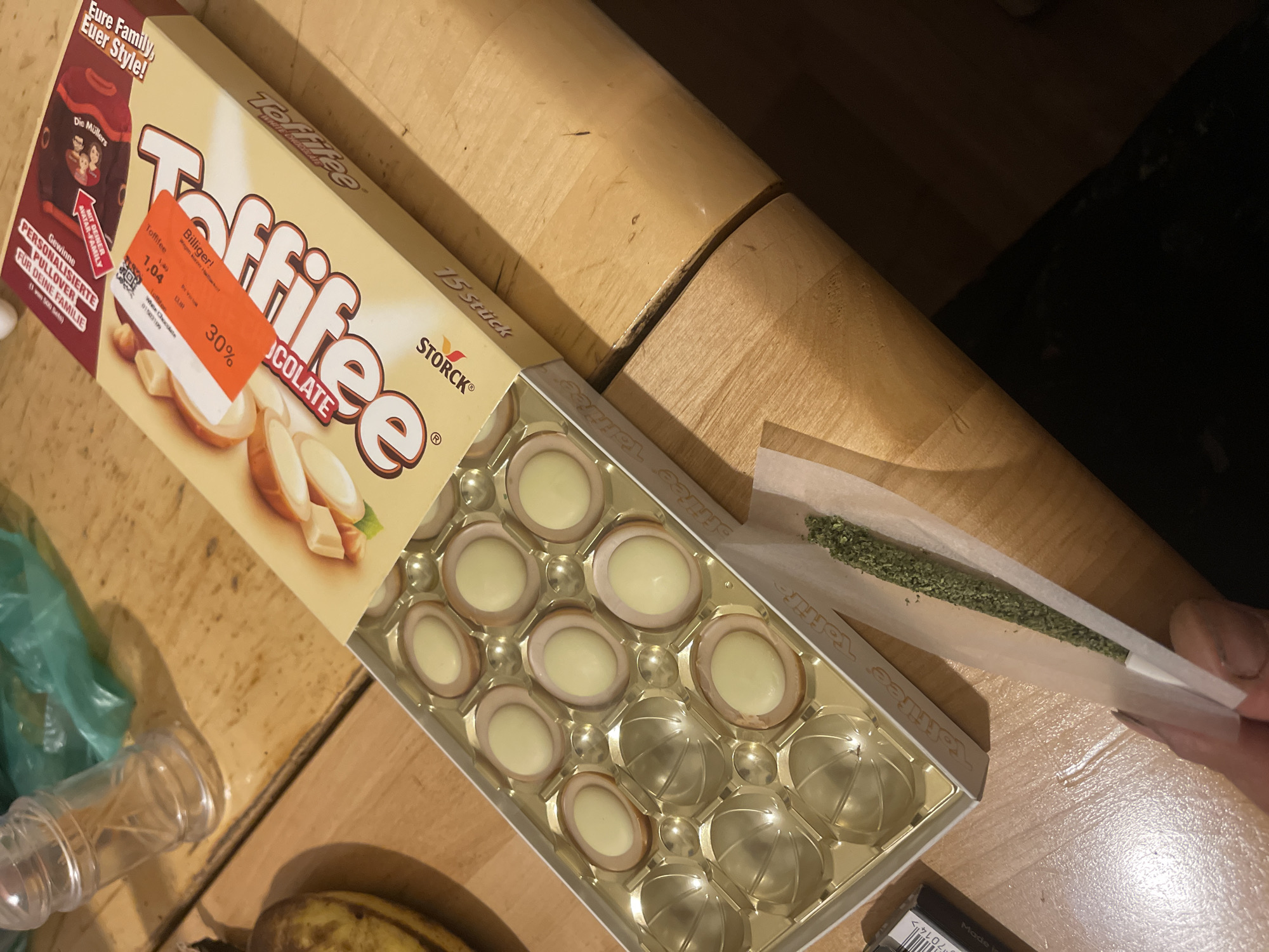 Here is a forensic description of the image:
The image shows an opened box of Toffifee white chocolate candies on a light brown wooden surface. The box is partially full, containing several candies, while some spaces are empty. Next to the box, a hand holds a cigarette filled with what appears to be a green, leafy substance. The background includes a plastic container, a banana, and an indistinct dark object. The scene is indoors, likely on a table. A promotional sticker with a discount percentage on the price of the Toffifee is visible on the package. The brand name "STORCK" is also visible on the box.