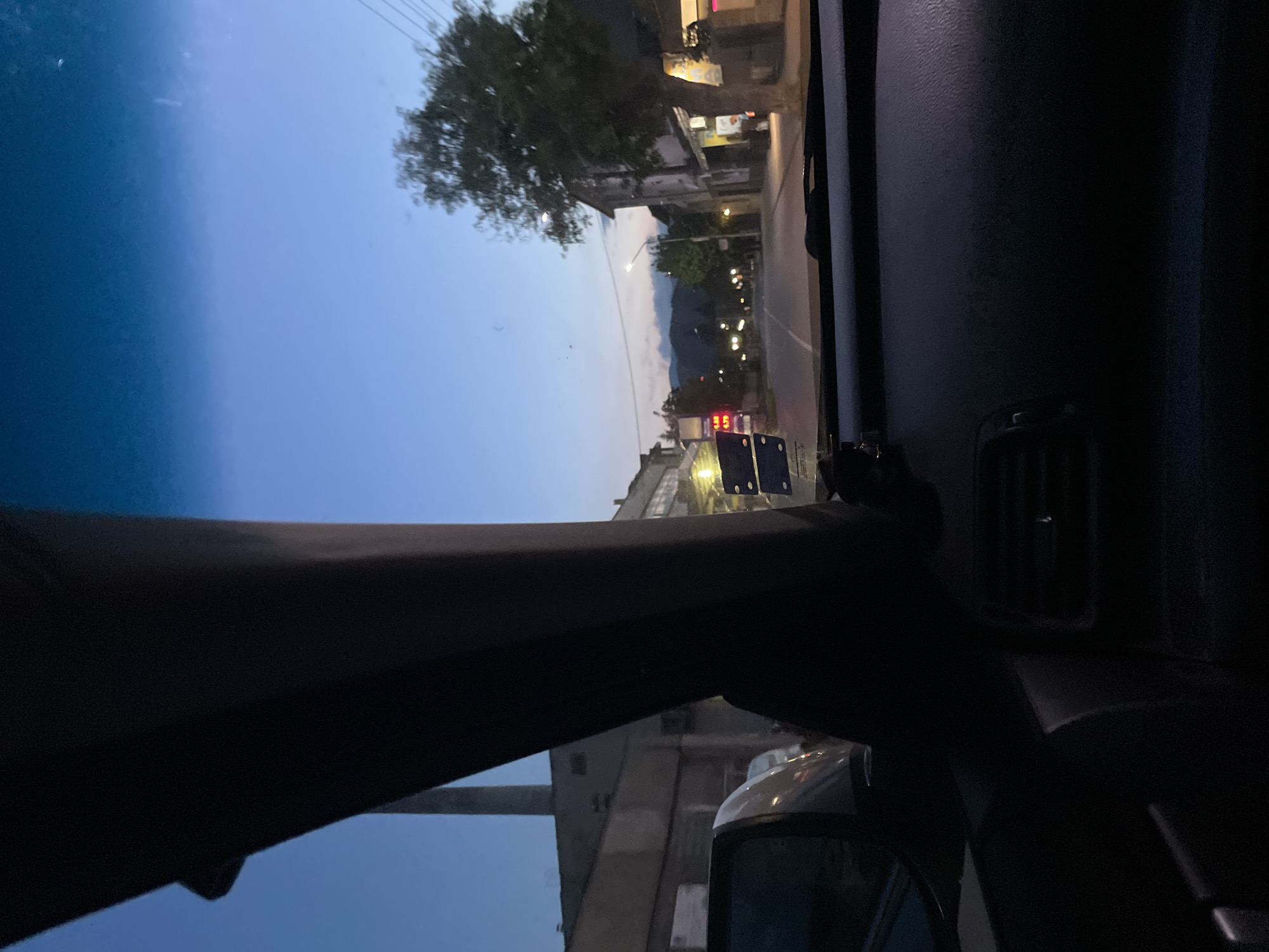 The image shows a street view in Zlatograd, Bulgaria, taken from inside a vehicle. It's evening, and the sky is a gradient of blue. The street is lined with buildings. There is a mountain range visible in the background. Streetlights illuminate the road. Part of the vehicle's interior, including the dashboard, window frame, and side mirror, is visible.