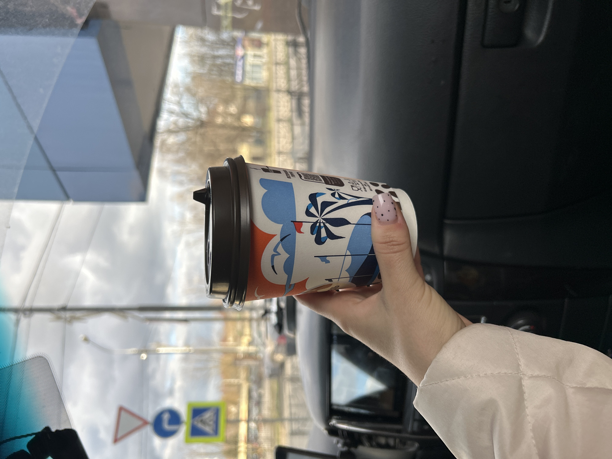 A woman's hand holds a coffee cup in a car. The cup has a brown lid and orange and blue graphic design. The woman's nail polish has a white base with black dots. The car's dashboard is black. Through the windshield, signs and buildings are visible indicating the location is in Klin, Russia. The sky is cloudy.