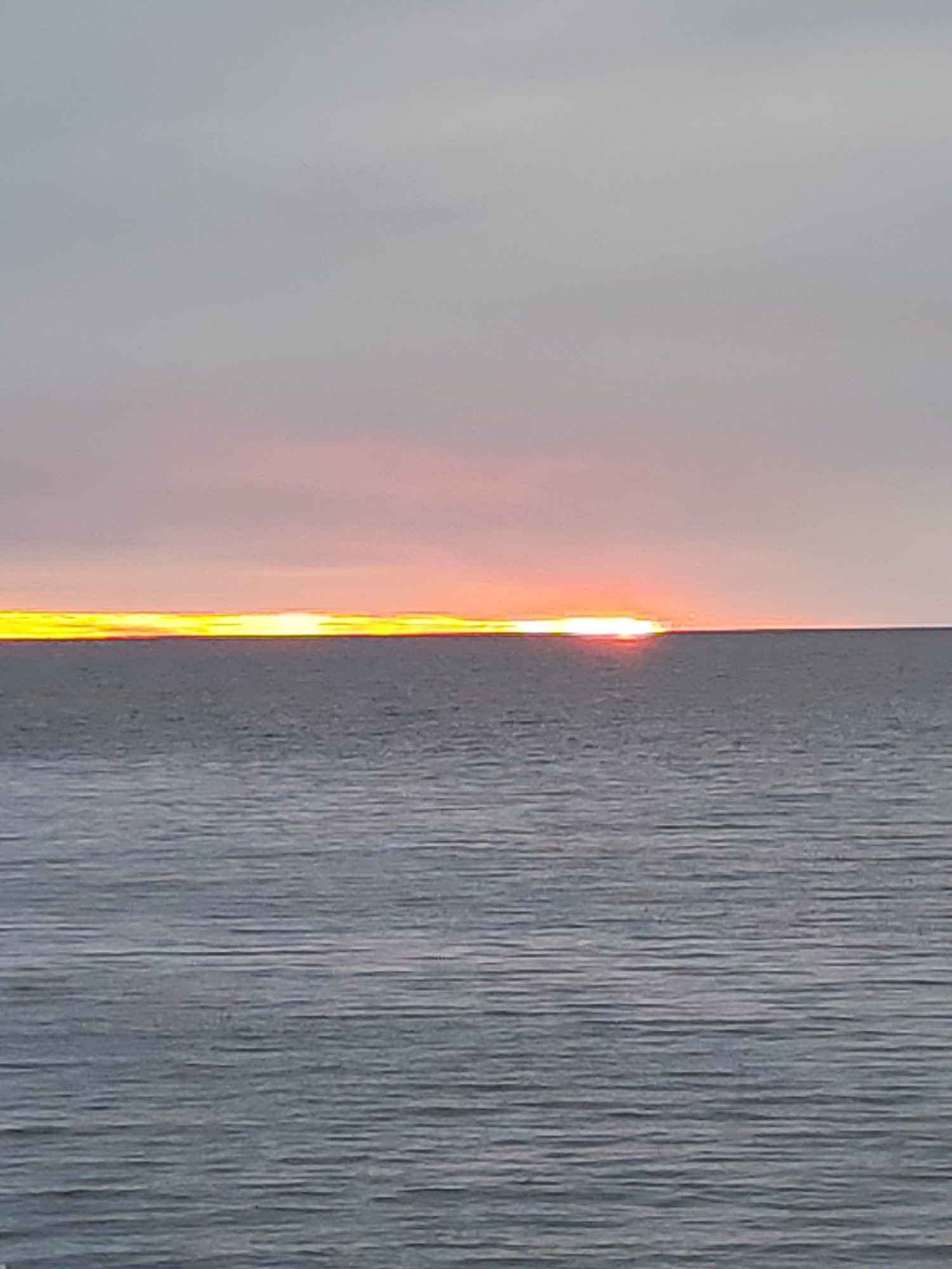 The image shows a sunset over water. The sky is a pale gray with a band of orange and yellow at the horizon, where the sun is setting. The water is a dark gray with subtle ripples. The image appears to be taken at sea level. There are no people or objects visible other than the sky and water. The location within the city of Shcheleiki, Russia is not discernible from the image.