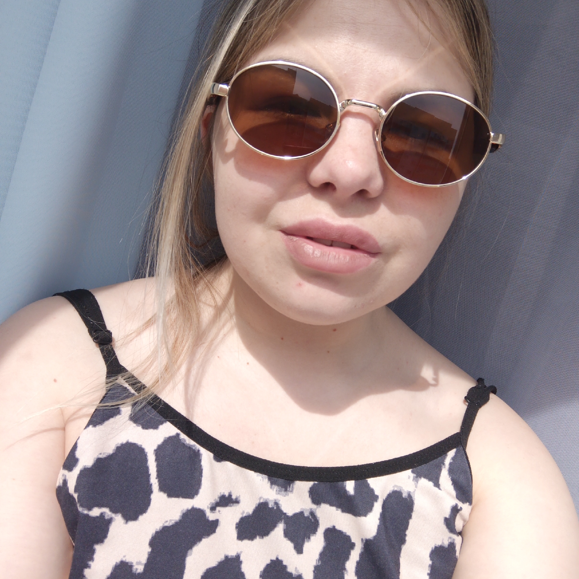 A young person, likely female, is featured in a close-up selfie, illuminated by bright sunlight. She has fair skin, light-colored hair, and is wearing round, gold-framed sunglasses with dark brown lenses, which reflect a bright, possibly indoor, environment or sky. Her lips are a soft pink.
She is wearing a tank top or camisole with a black and beige/cream abstract animal print design (resembling leopard or giraffe spots) and black trim and straps. One strap, visible on her right shoulder, has a small black adjuster or decorative element.
The background consists of a light blue/grey textured fabric, possibly a sunshade, curtain, or screen, suggesting an outdoor setting like a balcony or patio. Strong shadows are cast by the sunglasses on her face and by her hair/chin on her neck and shoulder, indicating direct and intense sunlight. The expression is neutral or slightly contemplative. No text is visible.