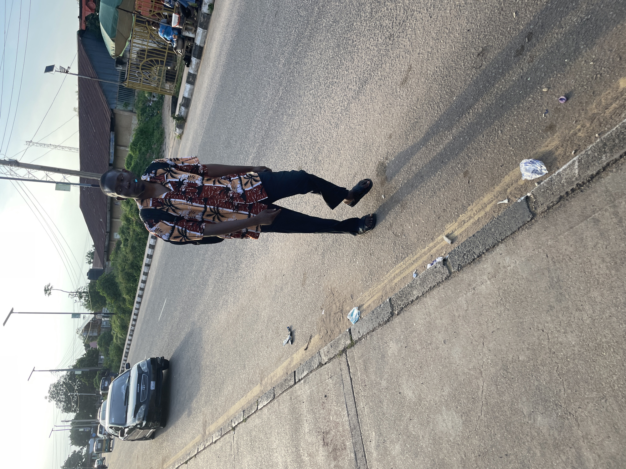 A man walks down a paved road, his shadow stretching long in the sunlight. He wears a short-sleeved shirt with a tropical print and dark pants. A backpack is slung over his shoulders. The road has a concrete curb and appears to be in a city setting. Litter is visible on the side of the road, including plastic bottles and wrappers. In the background, buildings and trees line the street, and vehicles, including a black sedan and what appear to be auto rickshaws, are driving on the road. The sky is overcast with a hint of blue. The overall impression is of a typical urban street scene in Jos, Nigeria, during daylight.