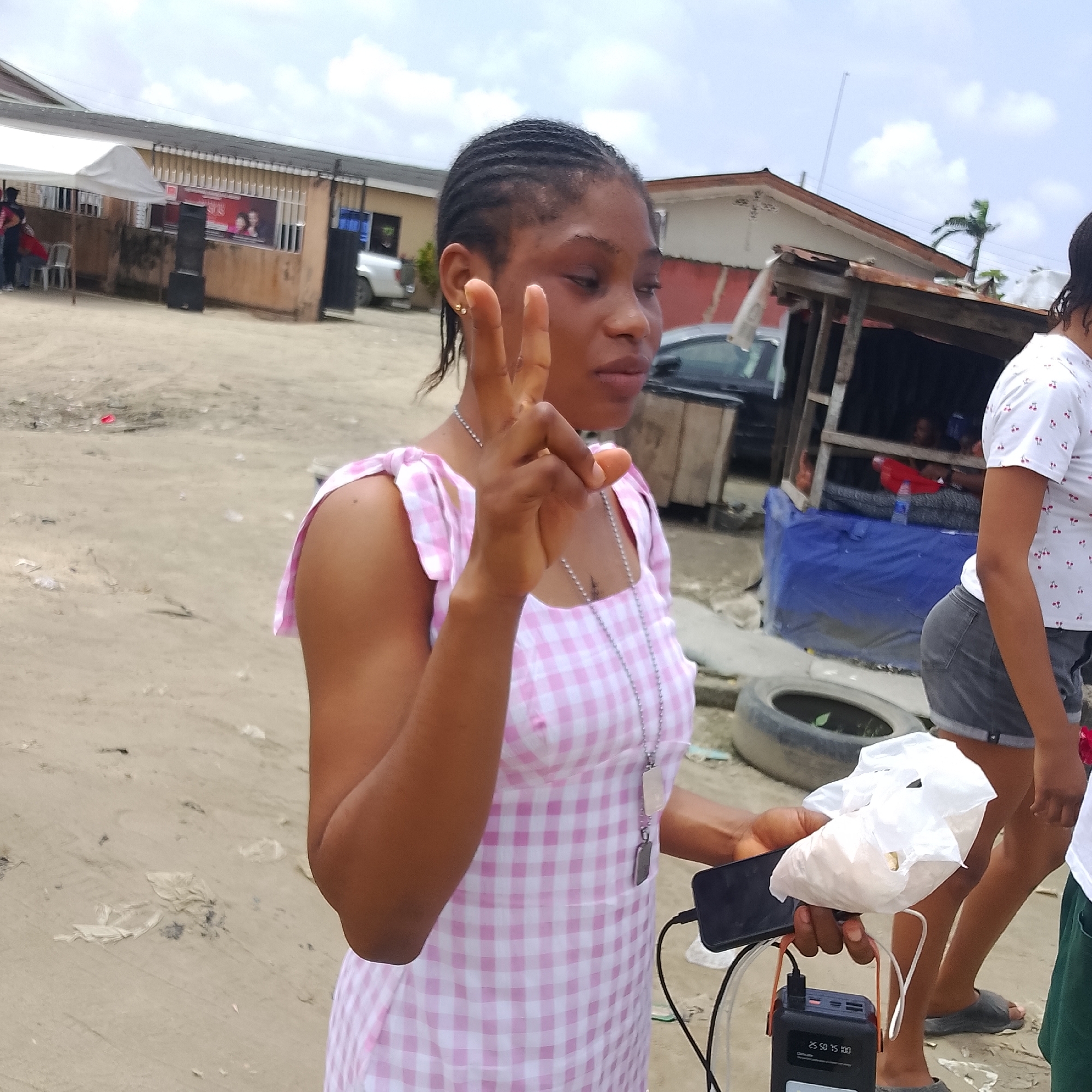 In Ado, Nigeria, a young woman with braided hair, wearing a pink and white checkered dress and a silver necklace, is shown in a three-quarter profile, holding a plastic bag of what appears to be food in her right hand and a smartphone in her left hand, which is connected to a portable power bank. Her left hand is raised, with two fingers extended in a peace sign. She is standing on a dirt ground. In the background, a partially constructed wooden structure covered with a blue tarp is visible, with other people and vehicles in the distance. The scene suggests an outdoor setting, possibly a market or a street, with a mix of commercial and residential elements. The lighting indicates it is daytime, with a cloudy sky.