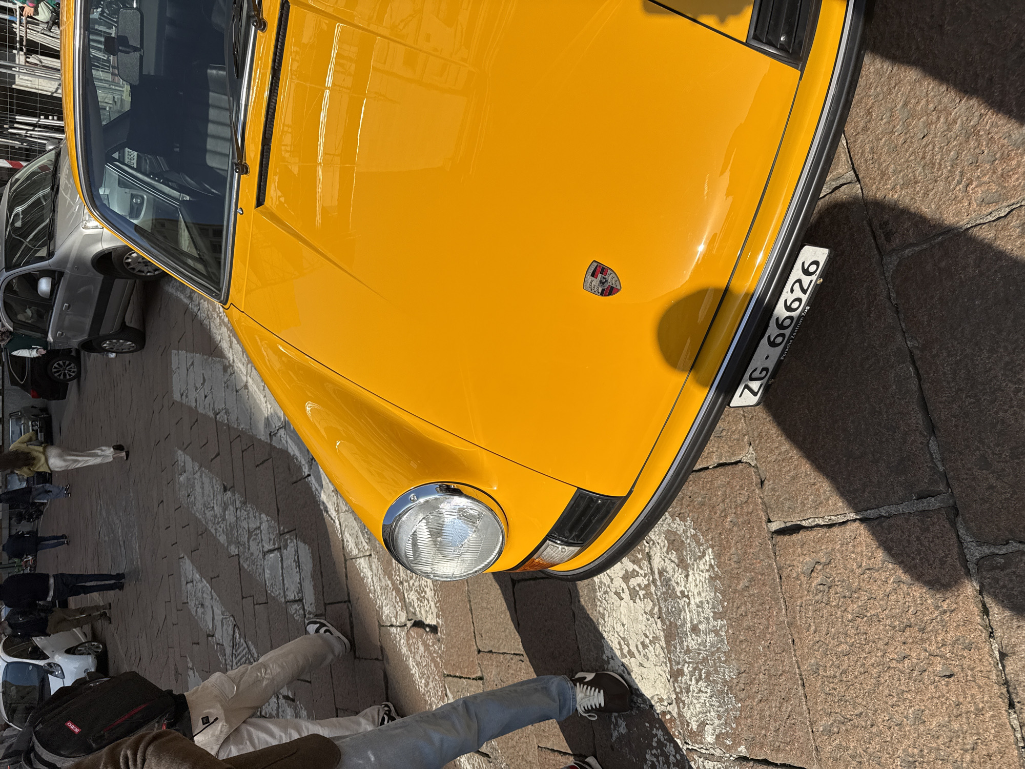 The image depicts an urban scene on a sunny day in Milan, Italy, characterized by a prominent yellow vintage Porsche 911 (likely a G-model) in the foreground. The car is angled towards the right, showcasing its front left side, including a circular chrome-bezeled headlight, an amber turn signal, and a black rubber bumper strip. The Porsche crest is visible on the center of the hood. Below the bumper, a white license plate with "ZG - 66626" and "Porsche Zentrum Zug" text is affixed.
The vehicle rests on a reddish-brown cobblestone street, which exhibits patches of weathered, lighter-colored material. Long, distinct shadows are cast from the left, indicating direct sunlight from a low angle.
In the mid-ground, several pedestrians are visible walking on the cobblestones. On the far left, partial views of two individuals' lower bodies are present: one wearing light blue jeans and brown sneakers, and another wearing light-colored cargo pants and black-and-white sneakers, carrying a black backpack with red accents. Further back, a person in a yellow top and white trousers is walking away from the viewer, along with several other figures in casual attire. A pedestrian crossing, marked by faded white stripes, is visible on the cobblestones behind the immediate foreground figures.
Behind the pedestrians and the yellow Porsche, other vehicles are parked or moving. A silver Fiat Panda is visible, partially obscured by the Porsche's windshield reflection, and other dark-colored cars are further down the street. To the right of the Fiat Panda, metal scaffolding or construction barriers are discernible. The scene suggests an active city street or square.