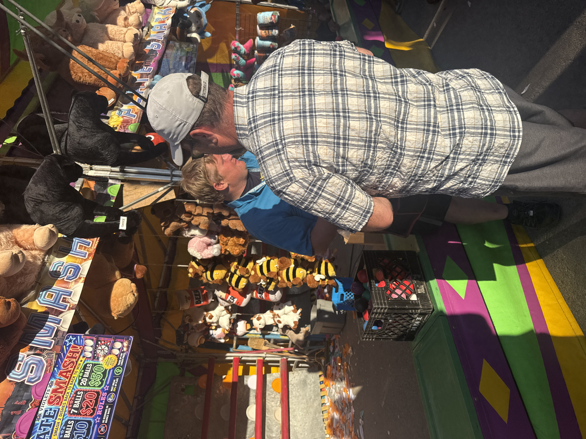 The image depicts a brightly lit outdoor carnival game booth, likely at a fair or amusement park. In the foreground, two individuals are visible. The person on the right, viewed from the back, wears a grey baseball cap and a white and blue plaid shirt with grey sweatpants. The person on the left, a younger male with blond hair, is wearing a blue t-shirt and appears to be interacting with the person in plaid. Behind them, the game booth is filled with plush toys, including several black panther-like stuffed animals and various other plush creatures hanging from the structure. A sign above the booth reads "SMASH!". In the lower left of the frame, a black plastic crate filled with colorful balls sits on a multi-colored wooden platform. The ground appears to be asphalt, and shadows from the booth and an unseen object are cast across the scene, indicating it is daytime and the sun is at an angle. There is debris on the ground in front of the booth. The overall impression is a typical scene at a summer fair. The location in Calgary, Canada is not specified within the image itself.