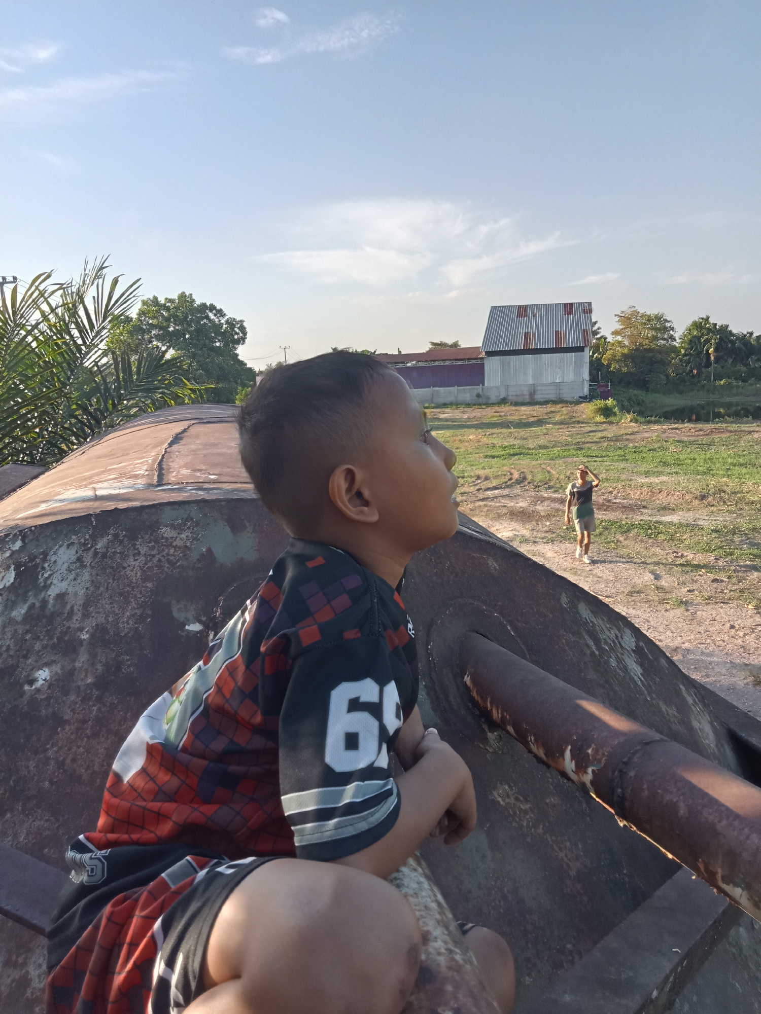 The image shows a young boy looking out to the right from atop a large, rusty, metallic object that appears to be part of a decommissioned tank. He is wearing a black jersey with red and orange plaid patterns and the number "6" prominently displayed in white. His expression is one of curiosity or contemplation.
In the background, a person in athletic shorts and a t-shirt is walking across a field of dry grass and dirt. To the left of the frame, palm fronds are visible, suggesting a tropical or subtropical environment. Behind the person in the field, there is a long, corrugated metal building with a red roof and a smaller, more modern-looking building next to it. The sky is a clear blue with a few wispy clouds, indicating it is daytime, likely late afternoon due to the warm, soft light casting shadows. The overall scene suggests an outdoor location, possibly in a rural or semi-abandoned area in Kualu, Indonesia. The rust and weathered appearance of the metallic object, coupled with the dry vegetation in the field, hints at a location that might be used for recreational purposes or is simply an overlooked part of the landscape.