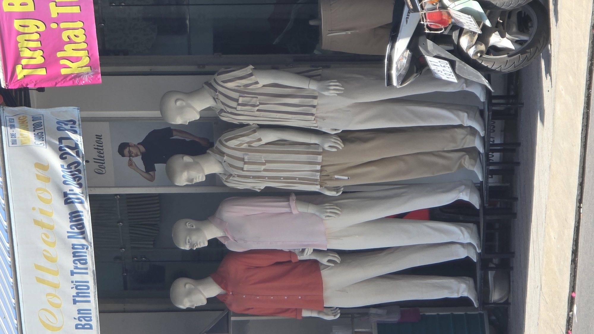 Here is a forensic description of the image:
This image appears to be taken outside of a clothing store, identified by a sign in Vietnamese that includes the words "Collection" and "Bán Thời Trang Nam" (Men's Fashion). The store has a glass window display showcasing several mannequins, each wearing different outfits. From top to bottom, these include two mannequins in striped short-sleeved shirts and light-colored pants, one in a light pink shirt and white pants, and one in a red shirt and white pants. A sign hanging near the entrance is bright pink with yellow text, partly obscured but appearing to read "Tưng Khai Tr". Above the mannequins, a motorcycle is parked near the storefront. The store is located in an area with sidewalks and other businesses visible in the background, which would put this place near the city of Nha Trang Ward, Vietnam.