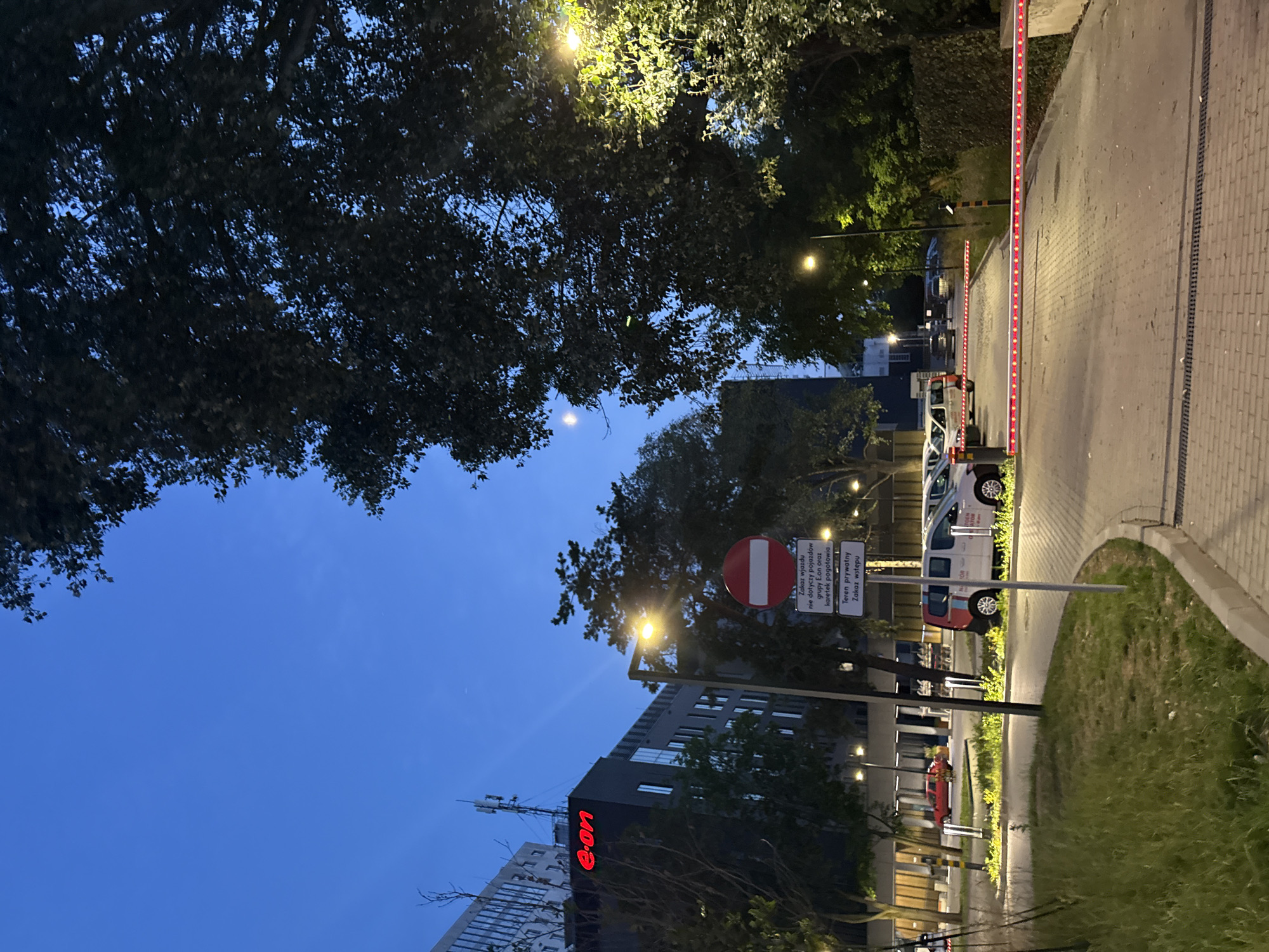 A street-level, vertical shot captures a city scene at dusk or dawn. The sky is a deep, clear blue. On the left, a modern, dark building with illuminated windows and a red "e-on" logo stands tall. Trees with dark foliage are scattered throughout the scene, some with branches reaching towards the sky. A lamppost with a bright, warm light illuminates the foreground. A circular red and white "no entry" sign is mounted on a pole, with a smaller rectangular sign below it. Further down the road, a white car with red and white lettering is parked, and a red car is visible in the distance. A section of a grey brick wall or hedge runs along the right side of the image. A retractable barrier with red LED lights is lowered across a paved driveway, blocking access. The ground is paved with light-colored interlocking bricks.