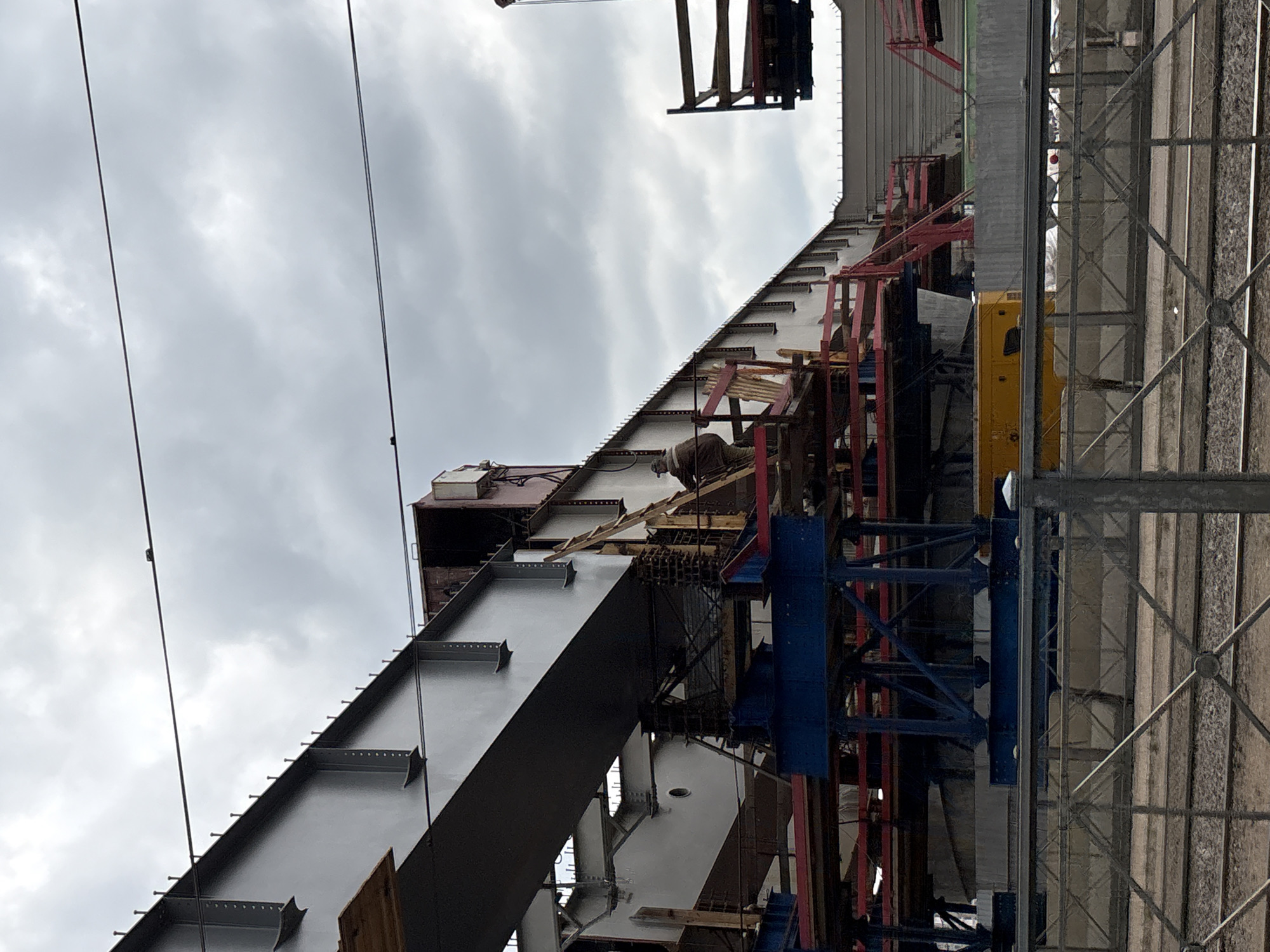 An overcast sky looms over a construction site where a large metal bridge section is being assembled. A lone worker, wearing a cap and a brown jacket, stands on a scaffolding platform to the right, appearing to be engaged in work on the structure. The bridge is supported by a complex network of blue and red scaffolding, with wooden planks and metal beams forming the infrastructure. In the foreground, a metal fence runs along train tracks. The scene depicts ongoing infrastructure development in an urban or semi-urban environment, given the presence of train tracks and what appears to be industrial architecture in the background. The location is identified as Unknown, Russia.