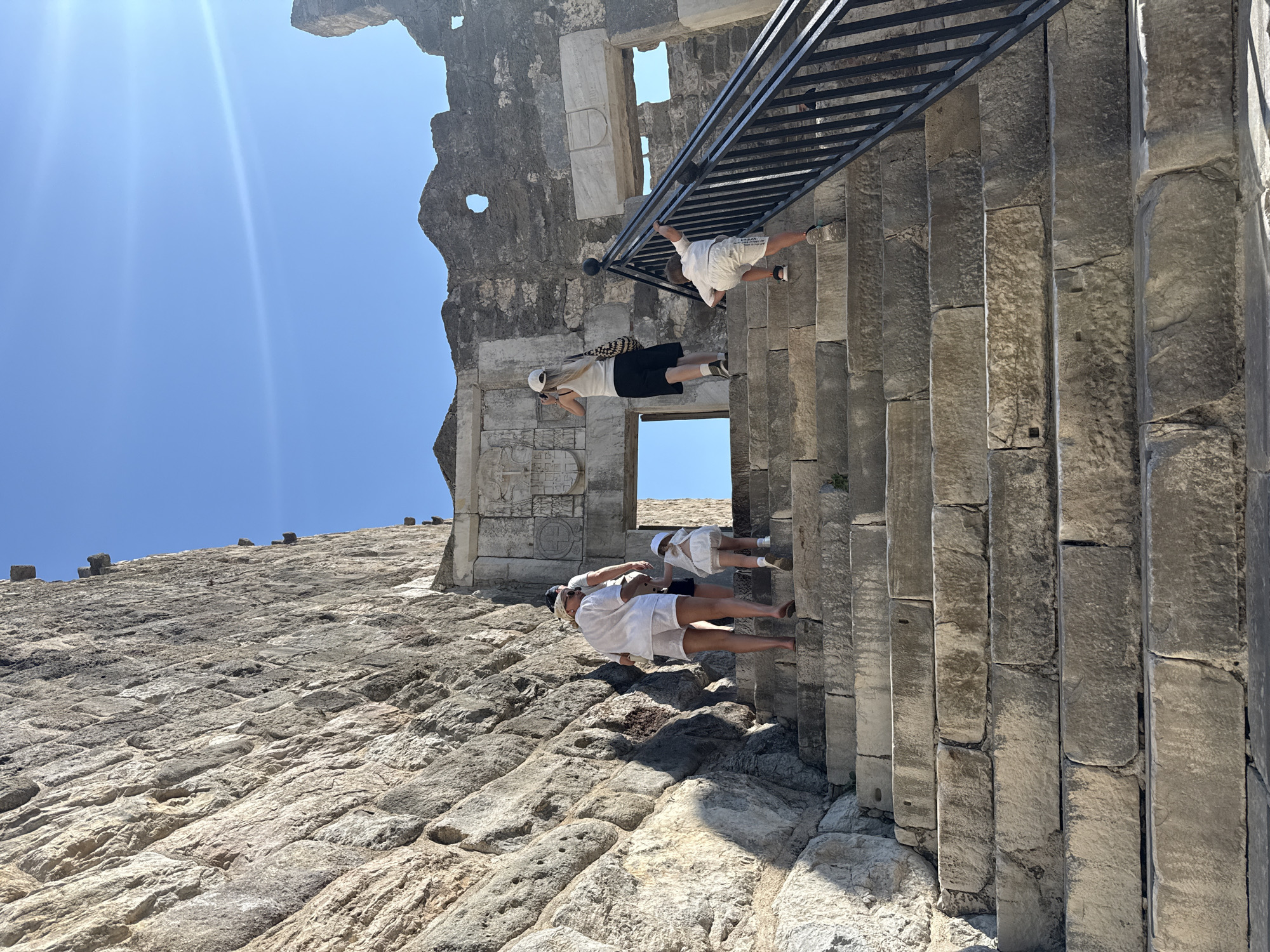 In Bodrum, Turkey, a family is ascending a stone staircase towards an ancient structure under a clear blue sky. A large, rough-hewn stone wall dominates the left side of the frame. The stairs are wide and made of massive, weathered stone blocks.
Several people are visible on the stairs. In the foreground, a young child in white shorts and a t-shirt is climbing on the right side, holding onto a black metal railing. Slightly behind and to the left, an adult wearing white shorts and a white top, with a white headscarf, walks up the stairs. To their left and slightly behind, another person in white is partially visible. Further up the stairs, another adult in a white tank top and black shorts is visible, holding a camera. A small child in a white dress is walking ahead of the adults.
Above the stairs, an arched opening reveals a bright sky. The structure appears to be ancient ruins, with stonework visible around the opening and on the right side, including some weathered carvings or crests above the archway. Sunlight casts shadows on the stairs and the wall, indicating a bright day. The overall scene depicts a moment of exploration and ascent within a historical setting.