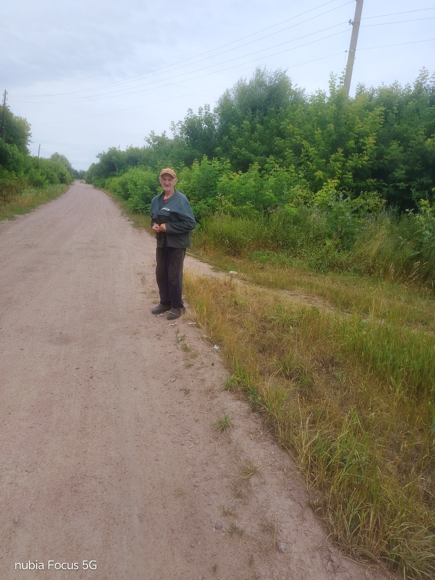 This is a photograph taken outdoors, likely in a rural area, possibly in Sestrynivka, Ukraine, given the context. The scene depicts a dirt road stretching into the distance, flanked by lush green trees and vegetation. The sky is overcast, suggesting a cloudy or possibly rainy day, and the time of day appears to be daytime.
The central subject of the image is an elderly man standing on the right side of the dirt road, facing the camera. He is wearing a light brown cap, a dark green jacket over a red shirt, and dark trousers. His posture is relaxed, with his hands clasped in front of him. He has a slight smile on his face, conveying a sense of mild contentment or perhaps a friendly acknowledgment of being photographed.
To the left of the man and stretching out to the horizon is the unpaved road, appearing to be a country lane or a path through a wooded area. The road surface is light brown and dusty, with scattered small stones. On the right side of the road, a thick growth of grass and various plants can be seen, some of which are dry and yellow, indicating possibly late summer or early autumn. Beyond the vegetation, more trees form a dense backdrop.
In the background, a utility pole with power lines is visible on the right side, suggesting the presence of infrastructure even in this seemingly remote location. The overall mood of the photograph is peaceful and serene, capturing a simple moment of a person in nature. The lower left corner of the image displays the text "nubia Focus 5G".
