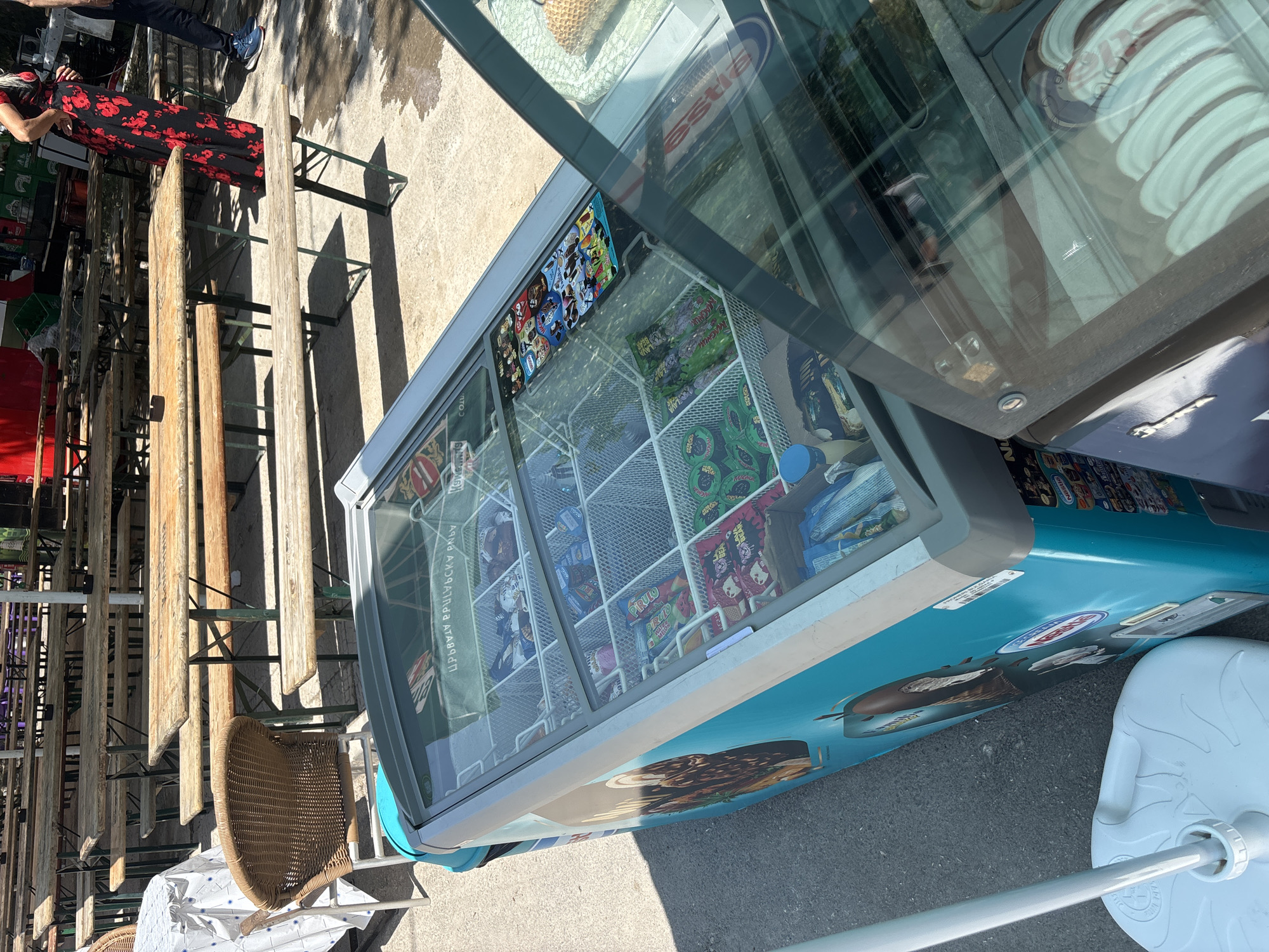 The image captures an outdoor commercial or recreational area in Kostievo, Bulgaria, under direct sunlight, indicated by distinct shadows. In the foreground, a light blue and grey ice cream freezer, bearing 'Nestle' branding on its side, is positioned centrally. Its clear glass lid displays multiple organized compartments containing various packaged ice cream and frozen confectionery products. Immediately to its right, a second, partially open display freezer contains multiple white, round tubs, consistent with scoopable ice cream. A white, circular base for an umbrella stand is located in the bottom-left corner.
The midground features several rows of weathered wooden picnic tables with integrated benches, arranged on a paved surface. To the left, a smaller, round table draped with a blue and white checkered tablecloth is situated next to a brown wicker chair.
In the background, a woman wearing a long black dress with red floral patterns stands between two benches on the right side, facing away from the camera. The lower body of a second individual, wearing blue athletic shoes and dark trousers, is partially visible at the far right edge. Green storage crates and a prominent red structure are discernible in the distant background, beyond the arrangement of tables.