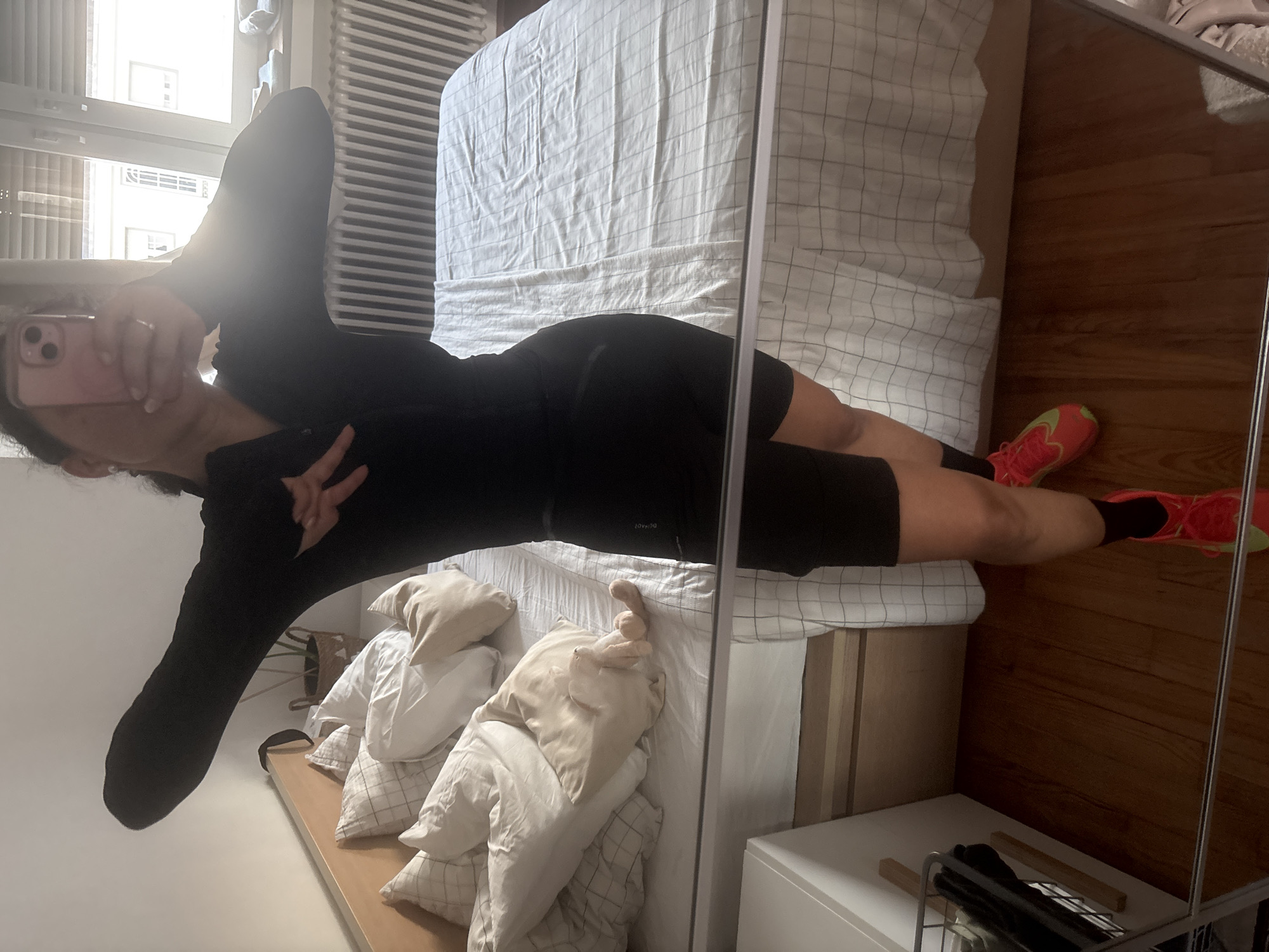 A full-body mirror selfie shows a person, likely female, standing in a bedroom. The person is wearing a fitted black long-sleeved top and black cycling shorts or athletic wear. They have dark hair and are holding a pink smartphone up to take the picture, partially obscuring their face. A ring is visible on one of their fingers. Their feet are adorned with bright red/orange and yellow/lime green athletic shoes with black socks. One hand is raised in a V-sign or peace gesture.
The background, seen through the mirror's reflection, indicates a brightly lit room. To the left, behind the person's head and upper body, a large window reveals buildings outside and a radiator below it, letting in natural light. To the right, a bed with a light-colored, possibly white or light grey, checkered duvet and pillows is visible. The bed frame appears to be made of light-colored wood. The floor is covered with dark wooden planks. In the foreground of the reflection, closer to the viewer, several light-colored pillows are stacked, and a white nightstand with a metal shoe rack or storage item is present. The time of day appears to be daytime due to the bright sunlight. No visible text is clearly discernible on any objects.
