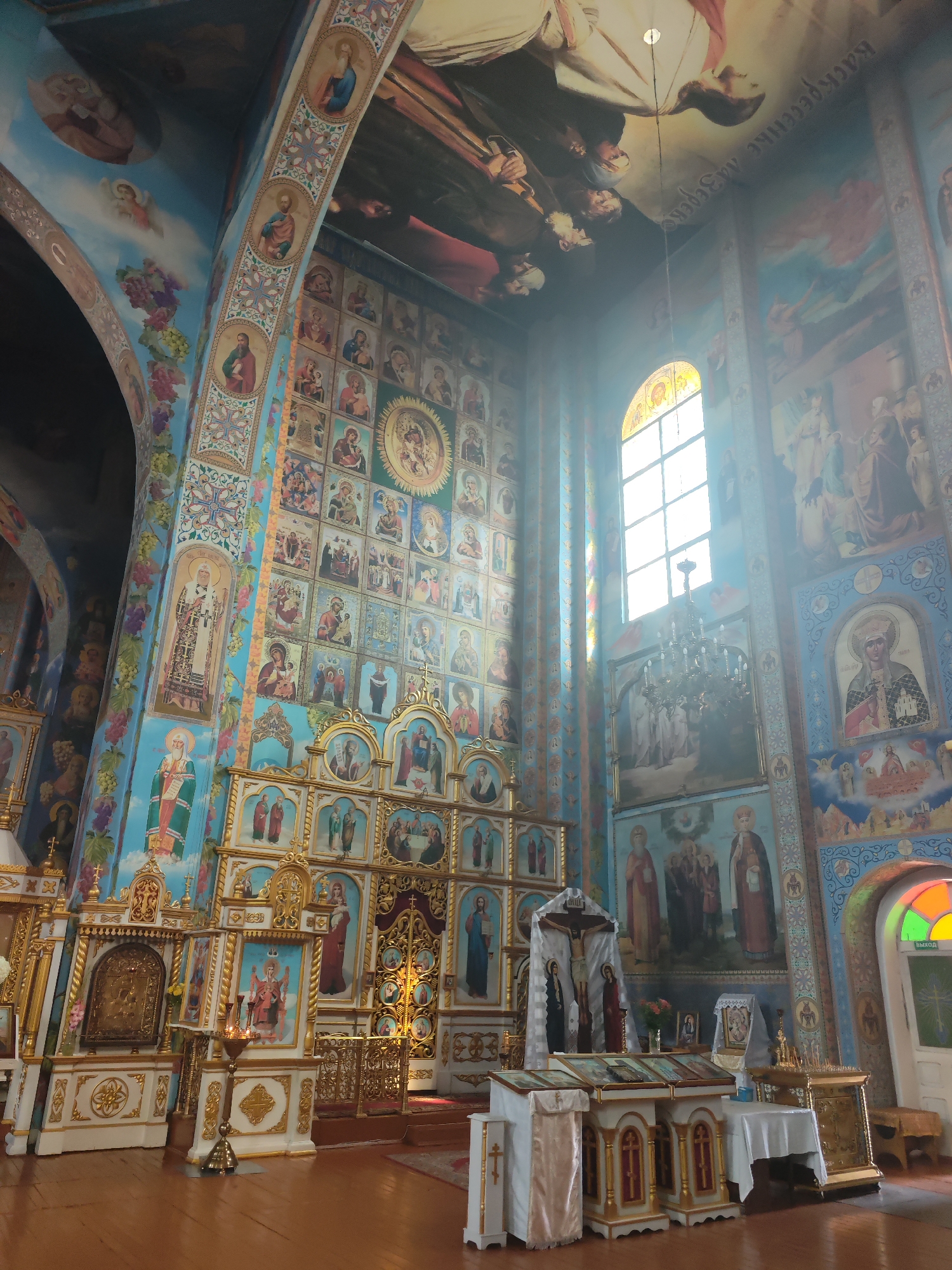 The interior of an Eastern Orthodox church is depicted, likely in Slonim, Belarus, given the style of religious art and architecture. The walls and ceiling are extensively decorated with religious frescoes and icons, primarily in blue, gold, and red hues. In the foreground, an ornate iconostasis, a wall of icons and religious images, stands prominently. Several tiered levels of icons are visible, many framed in gold. In front of the iconostasis are several white and gold altars with religious coverings and liturgical items, including candles and a cross with the crucified Christ figure draped in white cloth. To the right, a large arched window with stained glass in green, yellow, and orange sections illuminates the scene. A large chandelier hangs from the ceiling, casting light. The floor is made of polished wooden planks. The overall atmosphere is one of solemnity and religious devotion, with the bright, detailed artwork filling the entire space.