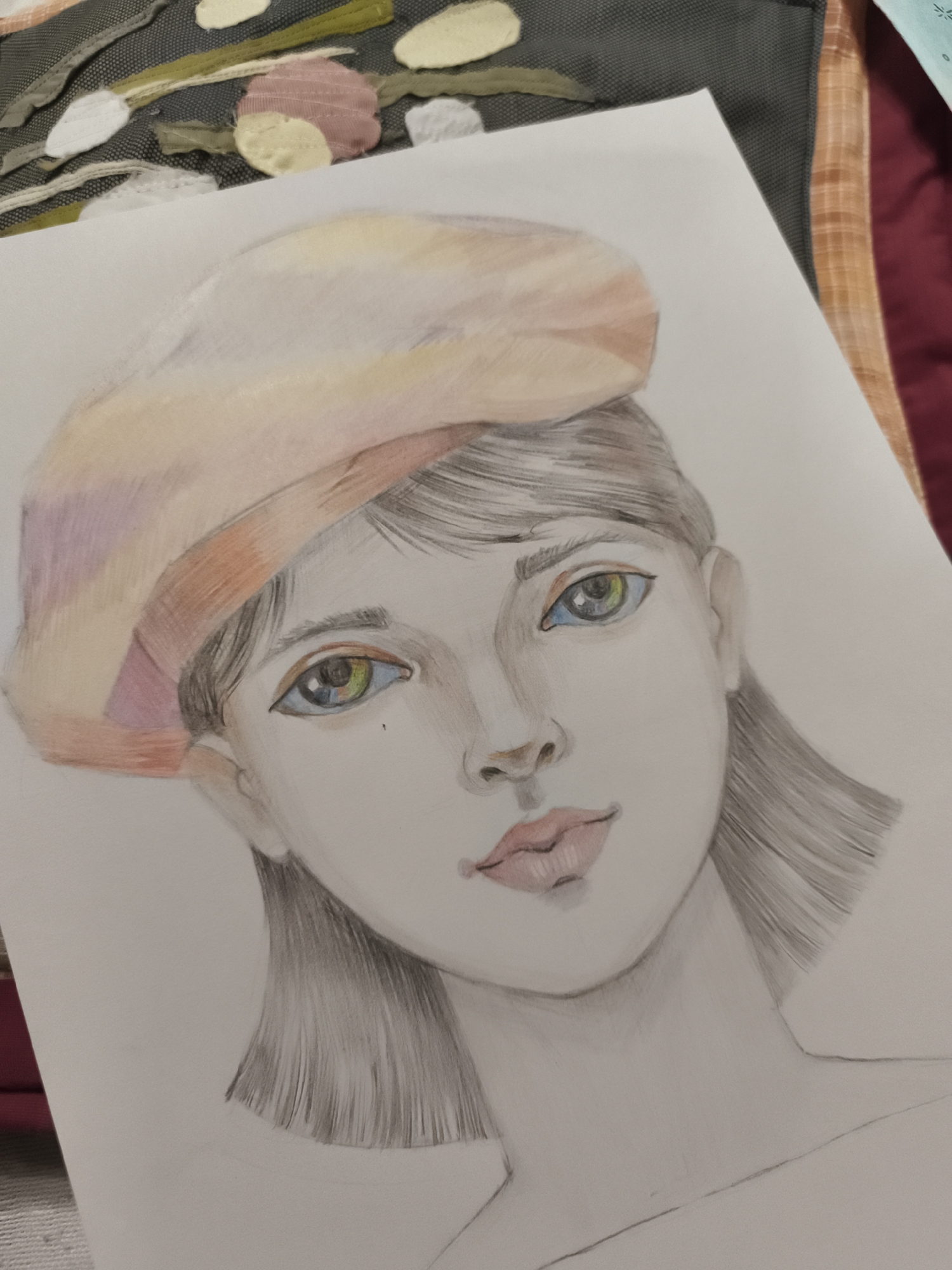 The image is a close-up, overhead shot of a drawing of a young woman wearing a beret. The drawing is rendered in colored pencils and graphite. The woman has short, dark hair, and her eyes are a striking mix of blue, green, and yellow. Her lips are painted a soft pink. The beret is a warm peach color with hints of pink and orange. The background of the drawing is plain white paper. The drawing is placed on top of a surface with a textured fabric, which appears to be part of a decorative textile with stitched shapes in various colors, including cream, pink, and green, arranged in a pattern. To the right, a sliver of a burgundy fabric with a plaid pattern is visible. There is no identifiable context related to the city of Saint-Denis, France, within the image itself.