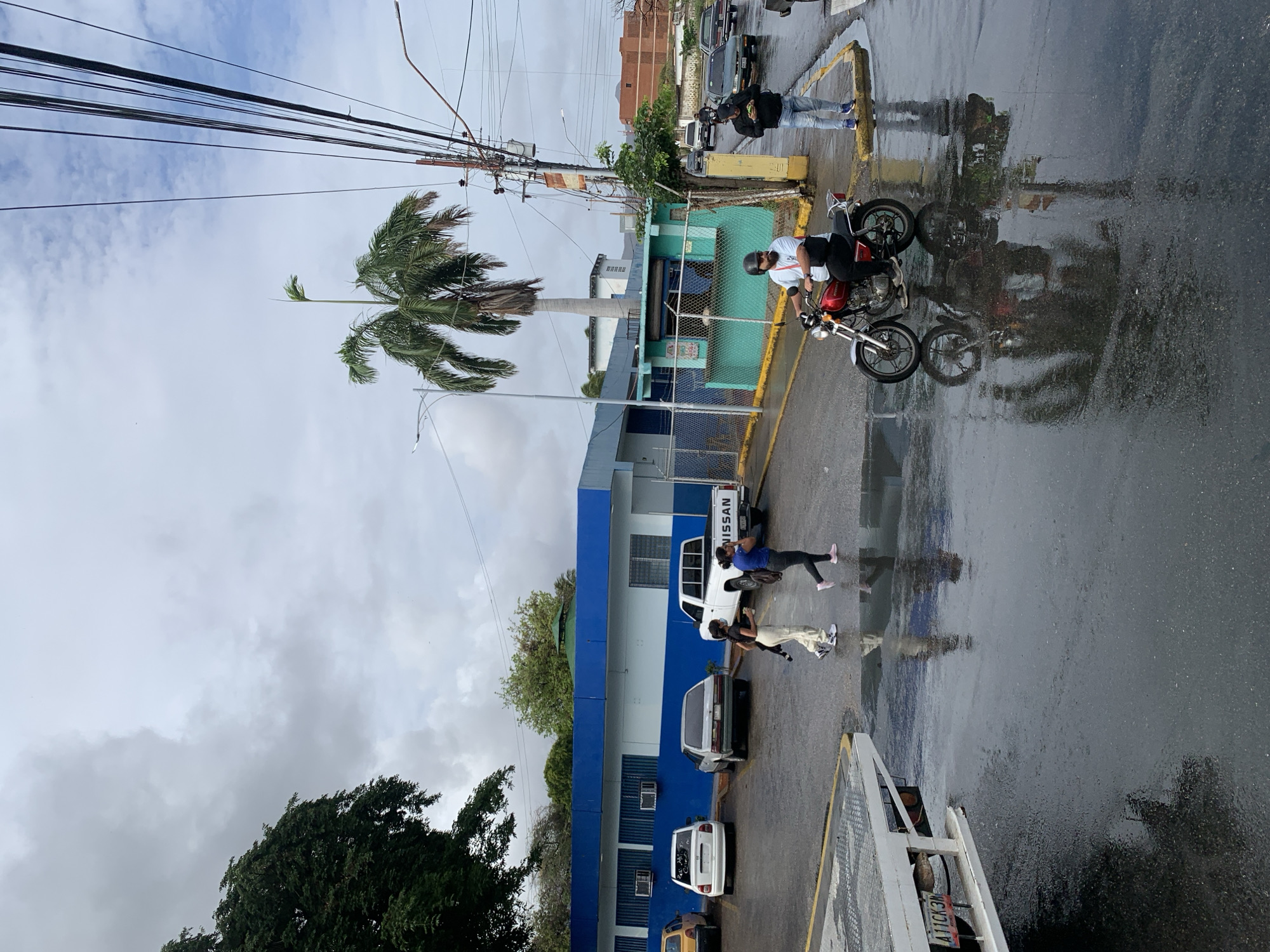 The image depicts a wet street scene in the city of Juan Griego, Venezuela, under an overcast sky. In the foreground, a white tow truck bed is visible on the left. Further into the scene, a man on a red motorcycle is riding away from the viewer. To his right, a man is standing on the sidewalk, looking at his phone. Two women are walking across the street, with one carrying a bag. In the background, several vehicles are parked, including a white Nissan pickup truck and a silver sedan. A blue and white building with air conditioning units is also visible, along with a small green building. A palm tree stands tall in the middle ground, and power lines crisscross the sky. The street is wet, with reflections of the surrounding objects visible on the surface.