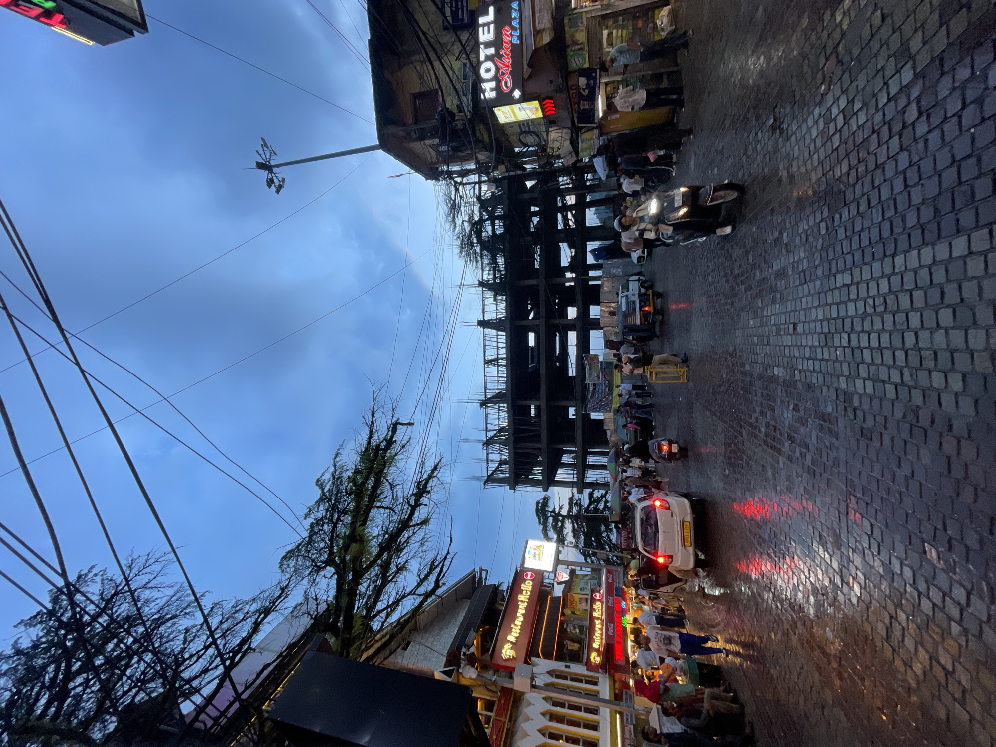 This image captures a bustling street scene in Bhagsu, India, likely during the twilight hours or after rain, given the wet, cobblestone pavement reflecting the ambient light. The street is lined with buildings, many of which appear to be shops and hotels, evidenced by signage such as "HOTEL Asian PLAZA" and "Restaurant Mello". A multi-story building under construction, with exposed steel framework, is prominently visible in the mid-ground.
The street is populated with a variety of people, some walking and others on vehicles. Several individuals are seen standing on the sidewalks, browsing or waiting. In the roadway, a white car with its brake lights illuminated is stopped, and a scooter with a rider is moving forward. Another scooter is also visible further down the street. An auto-rickshaw is also present. The sky is a cloudy blue, and overhead, a network of power lines crisscrosses the frame, a common sight in urban Indian settings. The overall impression is one of vibrant street life and commercial activity in a hilly or mountainous region, as suggested by the trees and the tiered construction of the buildings.
