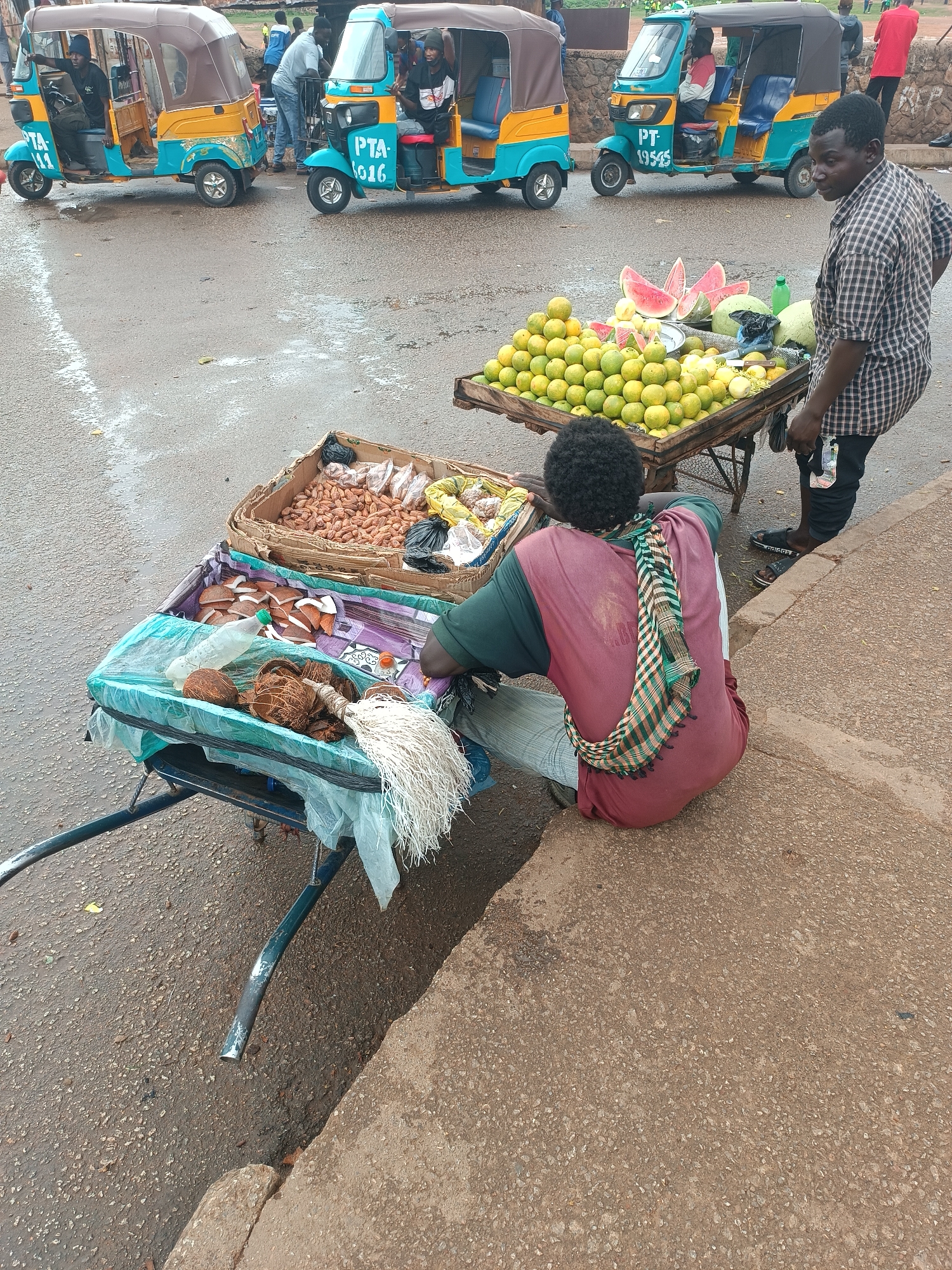 The image depicts a street scene in an unidentified urban area. Three auto-rickshaws are parked along the side of a road, all colored in light blue and yellow. Two men are engaged in commerce on the sidewalk next to the road. One man, wearing a plaid shirt and dark pants, stands beside a cart loaded with watermelons and citrus fruits. Another individual is sitting beside a cart filled with coconuts, a water bottle, and what appears to be nuts in a box. The setting suggests a local market or roadside vending area.