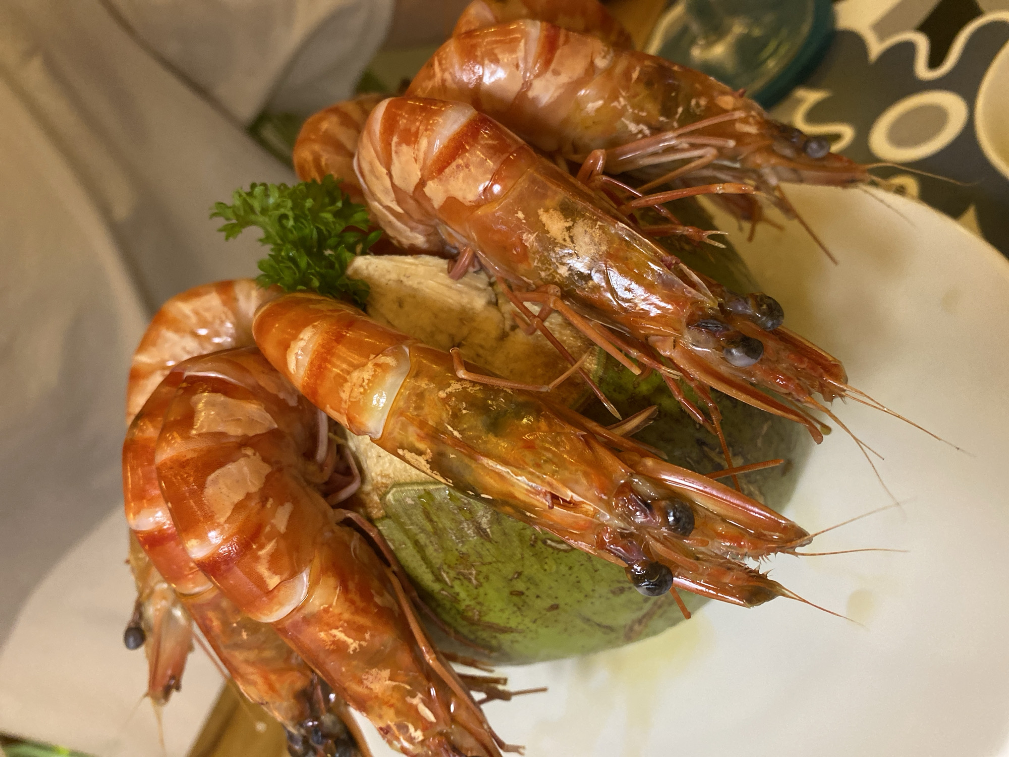 In Nha Trang Ward, Vietnam, a close-up shot captures a dish of large, cooked prawns arranged around a halved green coconut on a white plate. The prawns are bright orange with white stripes, their shells glistening. Some have their heads still attached, revealing dark eyes and thin antennae. A sprig of green parsley sits atop the coconut, adding a touch of color. The background is softly blurred, hinting at a casual dining setting.