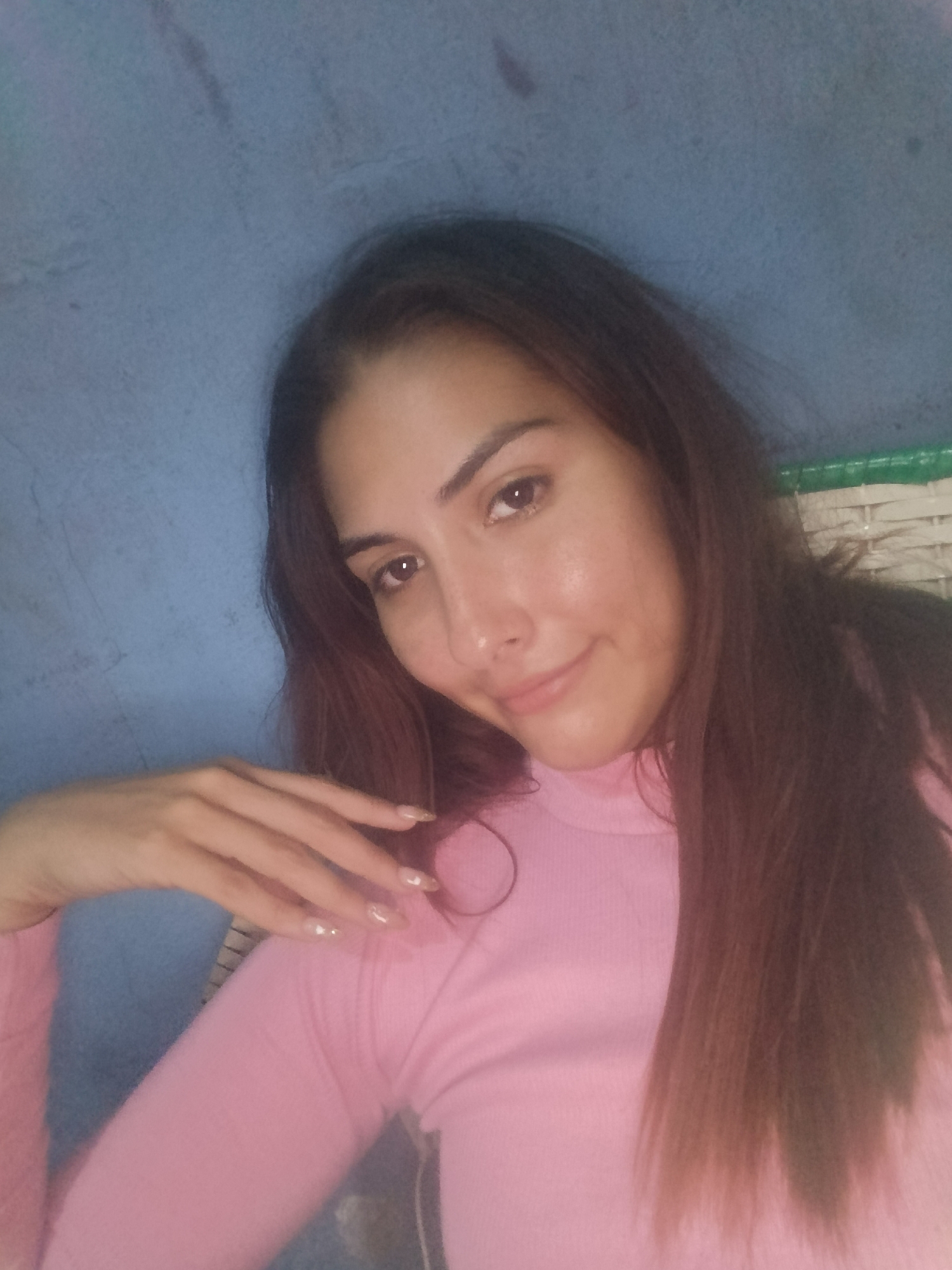 A woman with long brown hair and fair skin is posing for a photograph. She is wearing a pink turtleneck shirt. Her left hand is visible in the foreground. The background appears to be a textured blue wall. The location in Juan Griego, Venezuela, is not discernible from the image.