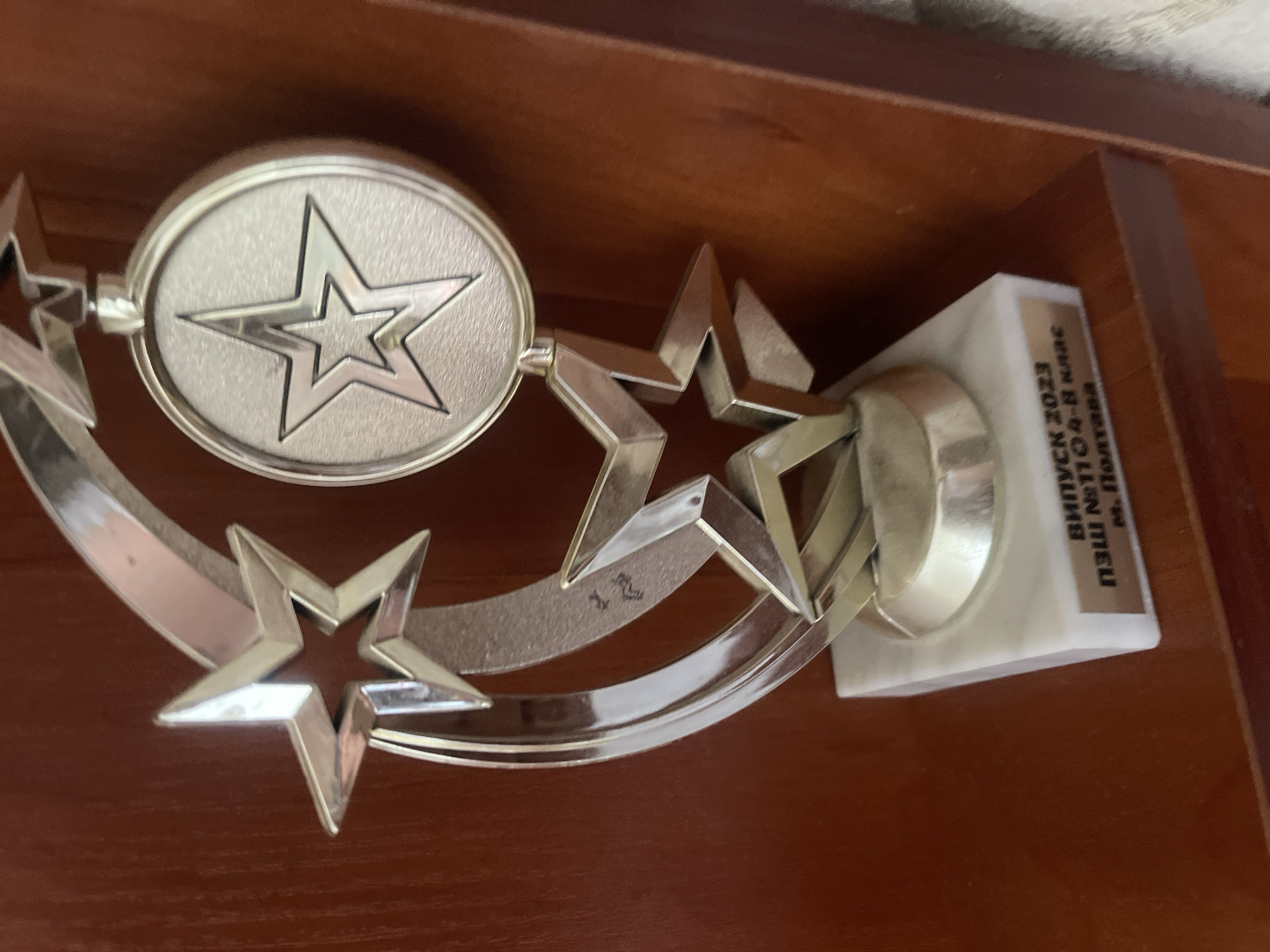 An award trophy with a star motif is displayed on a wooden shelf. The trophy features a crescent-shaped silver element adorned with three star shapes. One star is a pendant hanging from the crescent. The trophy rests on a white marble base with a golden plaque inscribed with text in Cyrillic. The text reads "ВЫПУСК 2023 ПЗШ №1104-8 класс М. Полтава". The background is a dark reddish-brown wooden surface.