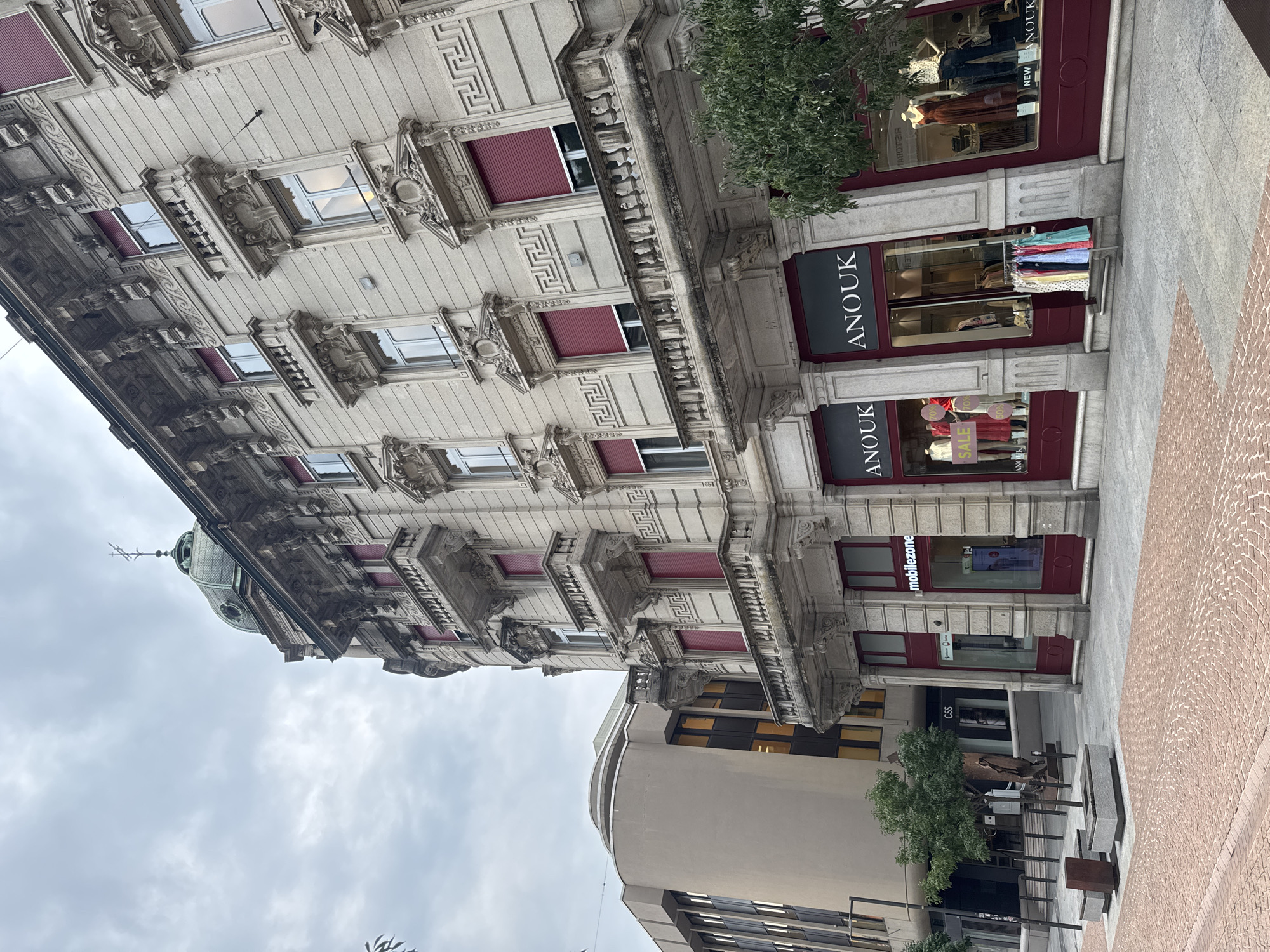 The image shows a street scene in Bellinzona, Switzerland, with a focus on the facade of a multi-story building. The architecture is ornate, featuring balconies with intricate railings and decorative carvings around the windows. Several windows have deep red shutters. On the ground floor, there are storefronts. One storefront displays the name "ANOUK" prominently, with mannequins inside dressed in clothing, and a rack of colorful dresses outside. Another storefront has a sign for "mobilezone" and appears to be a telecommunications store. To the left of the ornate building, there is a more modern building with a curved, beige facade and dark-tinted windows. The street is paved with a combination of gray stone and reddish-brown brickwork patterns. There are benches and planters on the sidewalk, along with some trees and a sculpture. The sky is overcast.