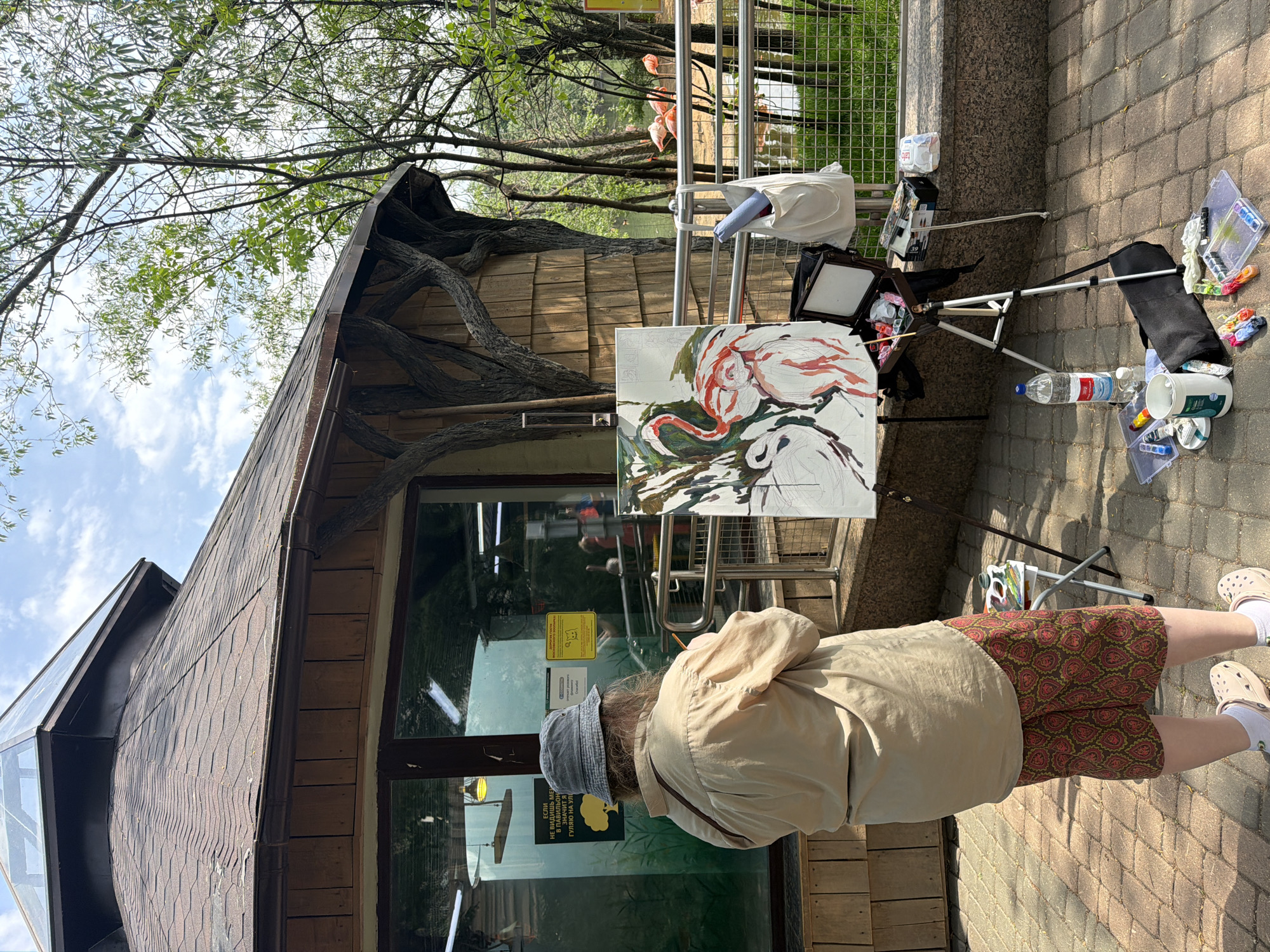 A person is painting on an easel outdoors next to a building with a textured, wood-like exterior. The person is wearing a beige jacket, patterned shorts, white socks, and beige shoes, and a gray hat. The painting on the easel depicts flamingos. Art supplies are visible around the easel on the brick ground. The building has windows with signage. In the background, a metal fence and trees are visible, along with several flamingos. Based on the architectural style and the signage language, the location appears to be in Moscow, Russia.
