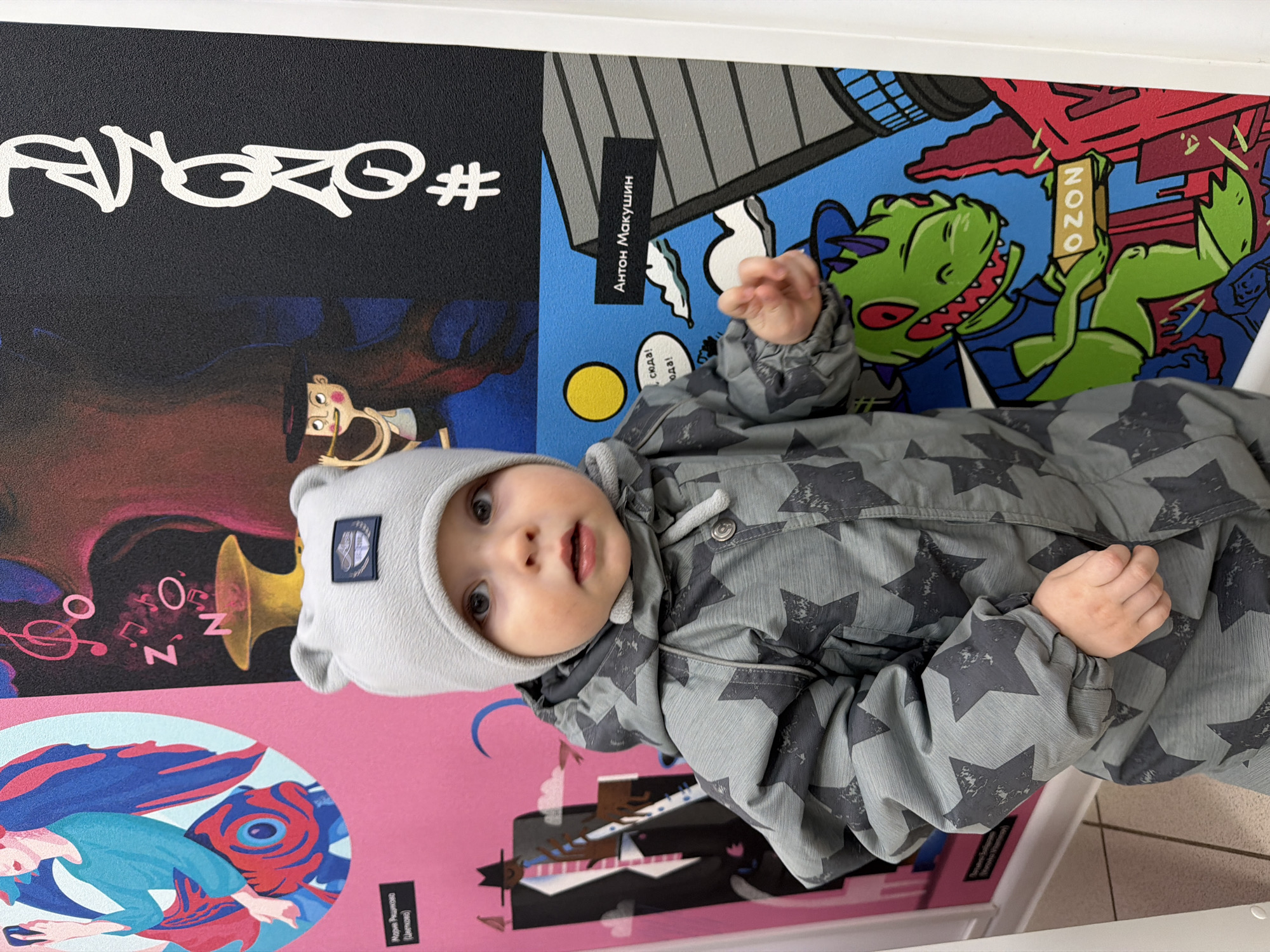 The media file features a baby, approximately 6-12 months old, dressed in a grey star-patterned snowsuit or jacket and a matching grey hat with ear-like details. The infant looks directly at the camera with wide, curious eyes and a slightly open mouth.
The baby is situated in an indoor setting against a colorful, multi-panel artistic mural or illustration. The background is divided into several sections:
1. **Top left:** A black panel displays white, stylized Cyrillic text identified by OCR as "AGRO#", with a partially visible figure below it.
2. **Top right:** A blue panel features a green cartoon dinosaur-like creature holding a sign with "OZON" written on it. A black label above the dinosaur reads "Антон Макушин" (Anton Makushin), and additional text says "сюда! сюда!" (here! here!).
3. **Bottom left:** A pink panel depicts a stylized blue and red aquatic or abstract figure, with the text "Мария Рещикова (Цветкова)" (Maria Reschikova (Tsvetkova)).
4. **Bottom middle/right:** A darker panel (black/dark red) shows musical notes, a golden trumpet, and the letters "ZON" arranged vertically, alongside another abstract figure.
The scene presents a cheerful and stimulating environment, with the baby appearing calm and inquisitive. The Cyrillic text and mention of "OZON" confirm the location context of Votkinsk, Russia.