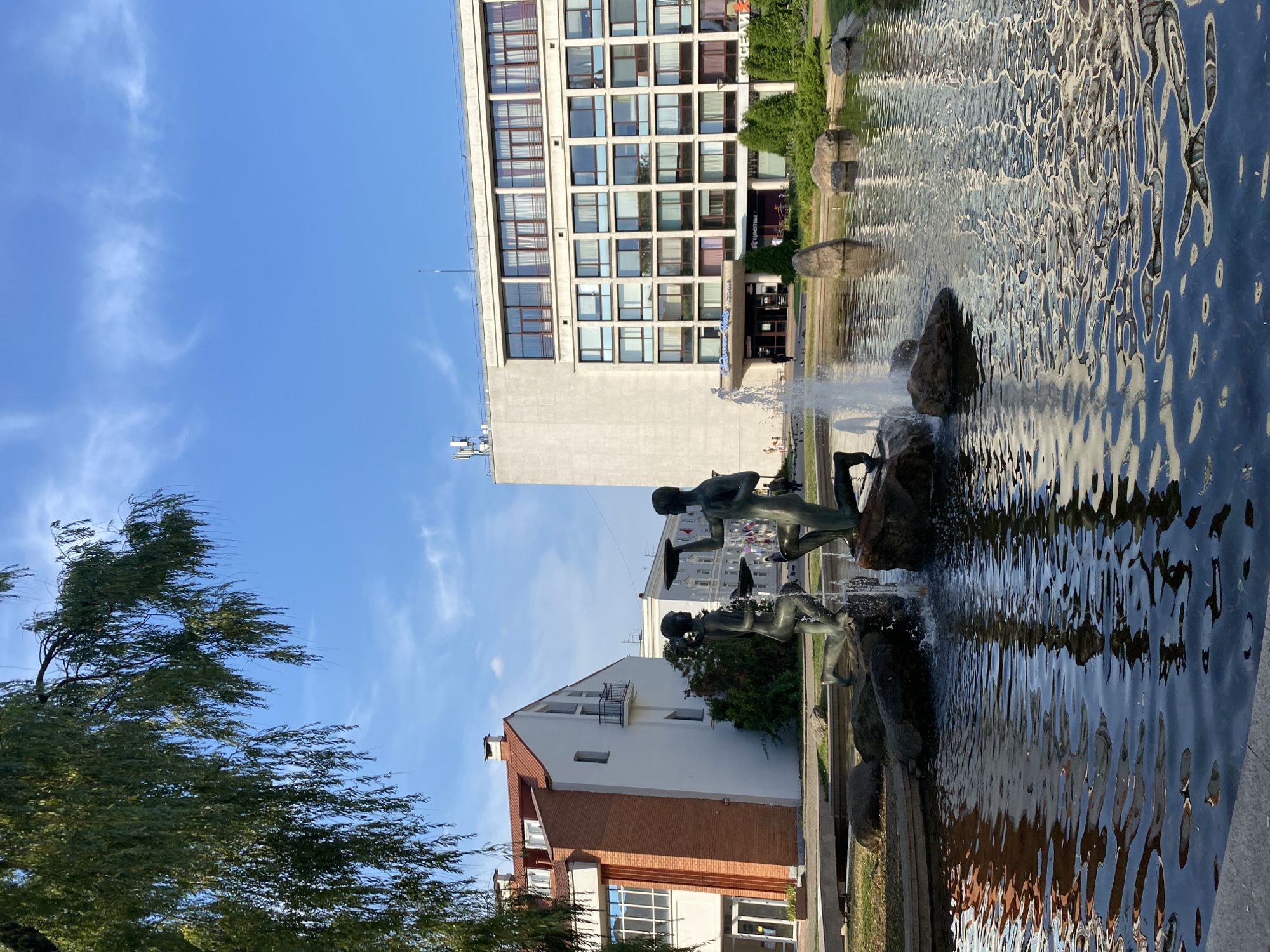 The image shows a public square in Minsk, Belarus, featuring a fountain with two bronze statues of human figures. The figures appear to be holding plates. The fountain is situated in a pond surrounded by rocks and greenery. In the background, there are several buildings, including a white building with a red-tiled roof and a larger, modern building with many windows. The sky is blue with some clouds. The area appears to be a pedestrian-friendly space, with people visible in the distance.