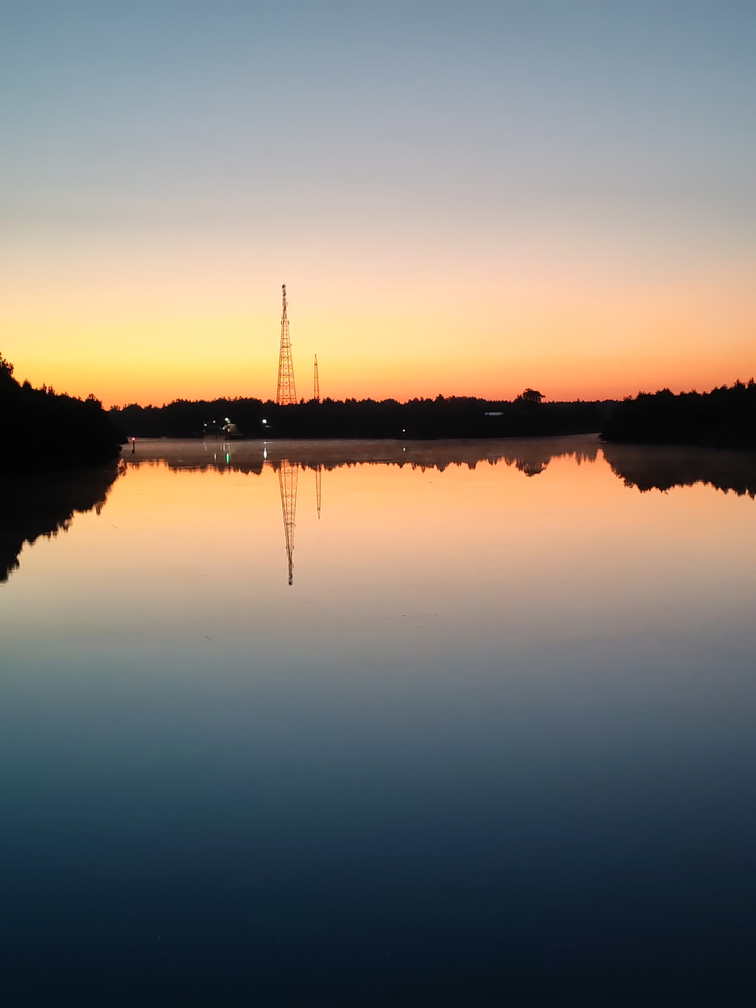 This is a landscape photograph taken during twilight, featuring a serene body of water reflecting the sky and surrounding landscape. The sky displays a gradient of colors, transitioning from a pale blue at the top to warm orange and yellow hues near the horizon, indicating either sunrise or sunset. Silhouetted against the colorful sky are trees forming a treeline along the edge of the water, and two tall, lattice-like towers. The water is calm, creating a near-perfect mirror image of the sky, trees, and towers. A faint mist hovers just above the water's surface, particularly noticeable near the treeline. The overall atmosphere is tranquil and peaceful. The image's metadata may contain location information, but based on visual cues alone, it depicts a natural or semi-natural environment at dusk or dawn. No people or animals are visible in the image.