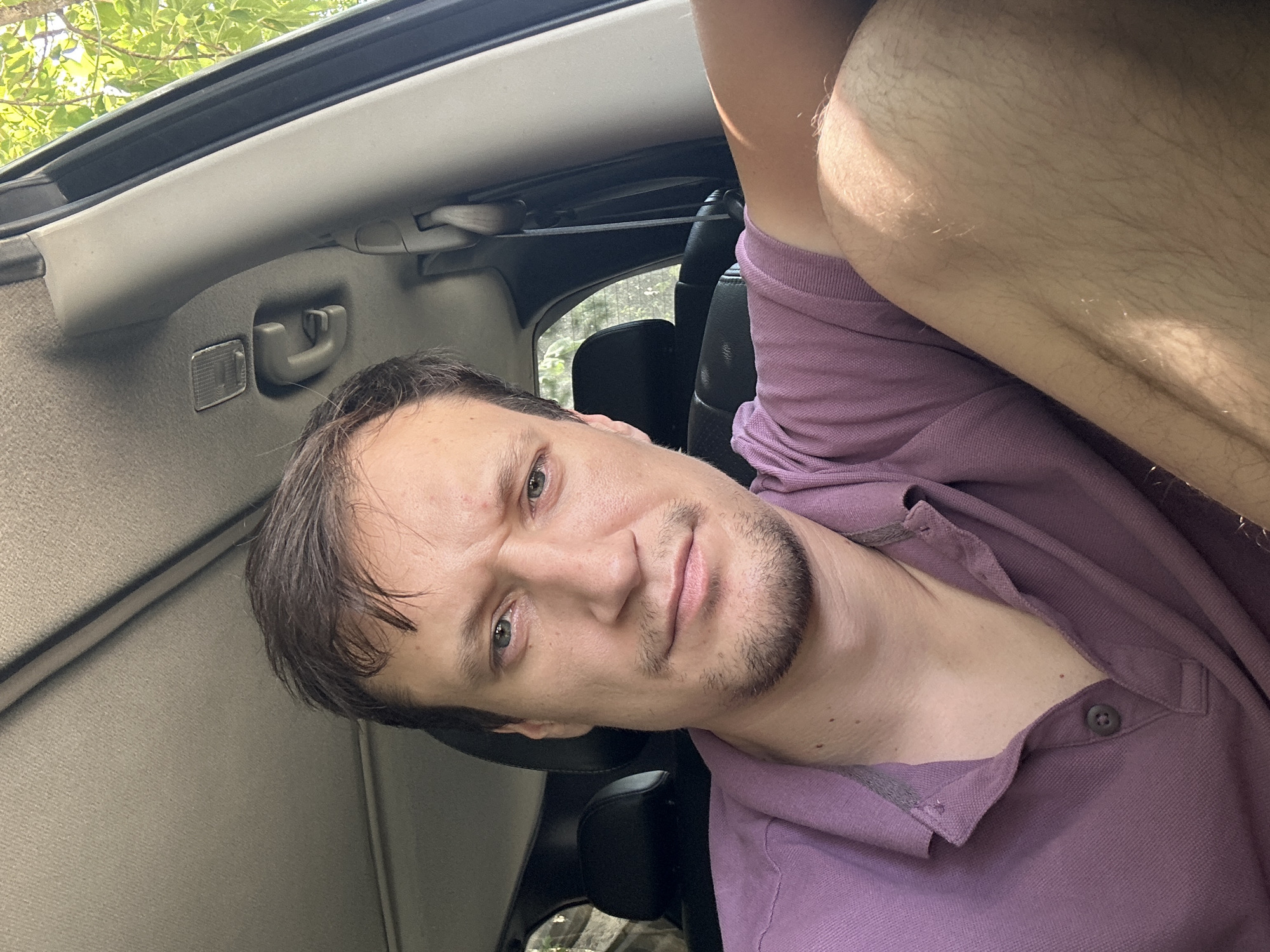 The image shows a close-up selfie of a man sitting in the passenger seat of a car, looking at the camera with a neutral expression. He has short, dark brown hair and light green eyes. He is wearing a purple polo shirt. The interior of the car is visible, including the headrest of the seat behind him and the car's ceiling. Sunlight is streaming in from the right side of the frame, illuminating his arm and leg. The background outside the car shows green foliage, suggesting a natural, possibly rural, setting. The location in Makiv, Ukraine, is not discernible from the visual information alone, as there are no specific landmarks or signs indicating this place.