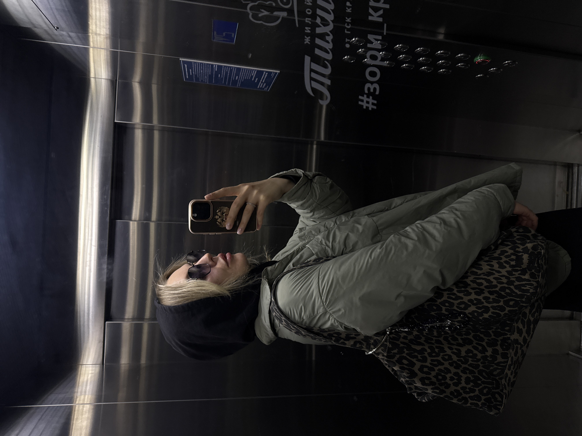 A person is taking a selfie in an elevator with mirrored walls. The person is wearing a sage green puffer jacket, a black hooded sweatshirt, and sunglasses. They are holding a cell phone with a decorative case. A leopard-print patterned bag is slung over their shoulder. The elevator's control panel is visible, along with signage in Russian. The city of Krasnoyarsk, Russia, is identified in the signage.