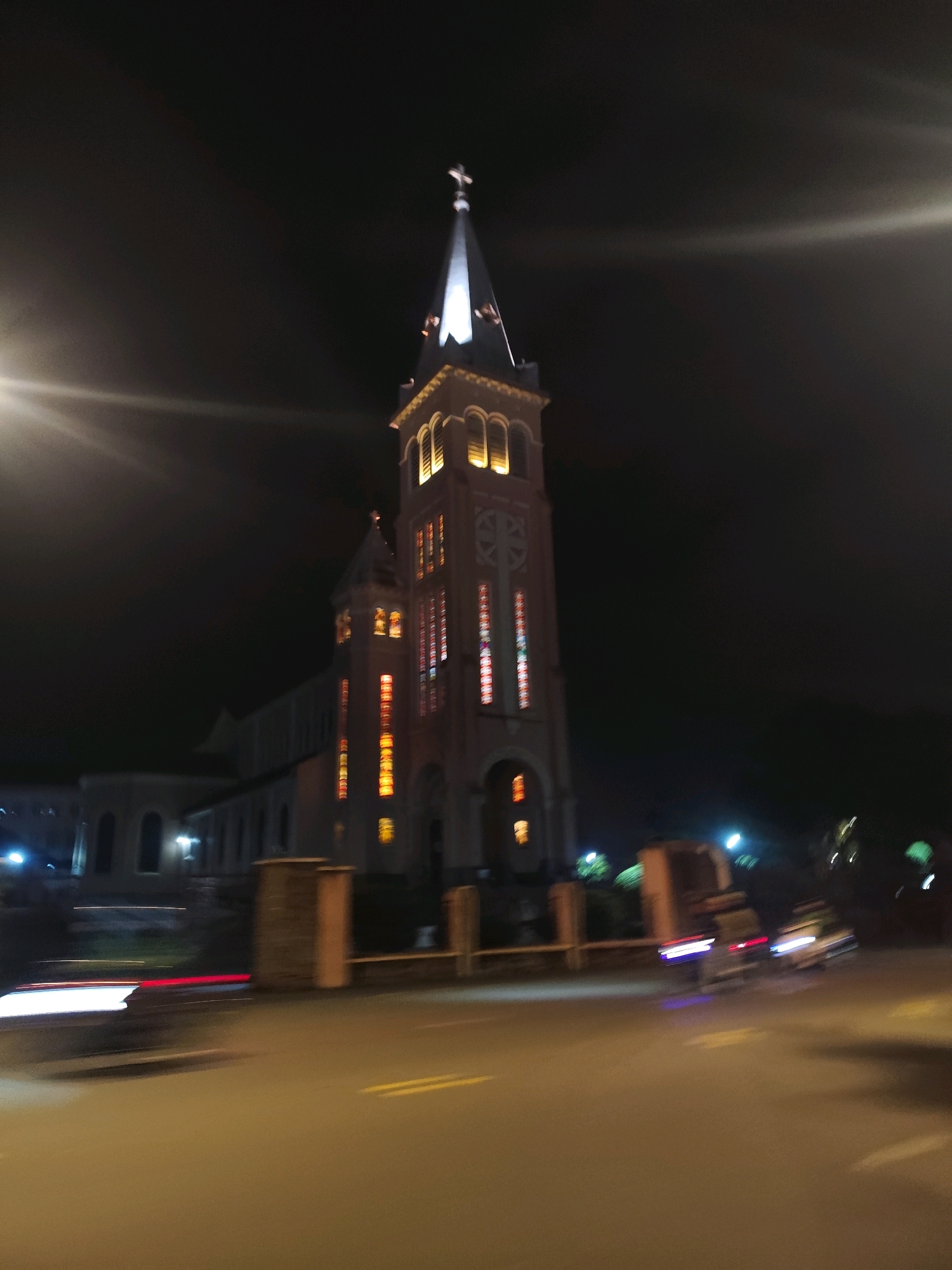 The image, taken at night in Da Lat, Vietnam, depicts a church with illuminated stained-glass windows and a tall spire topped with a cross. The church is a prominent architectural structure in the dimly lit urban environment. The street in front of the church shows motion blur from passing vehicles, indicated by streaks of white and red light. Two blurry figures are visible on motorcycles on the right side of the road. The overall atmosphere suggests a nighttime scene in a Vietnamese city, with the church serving as a central illuminated landmark.
