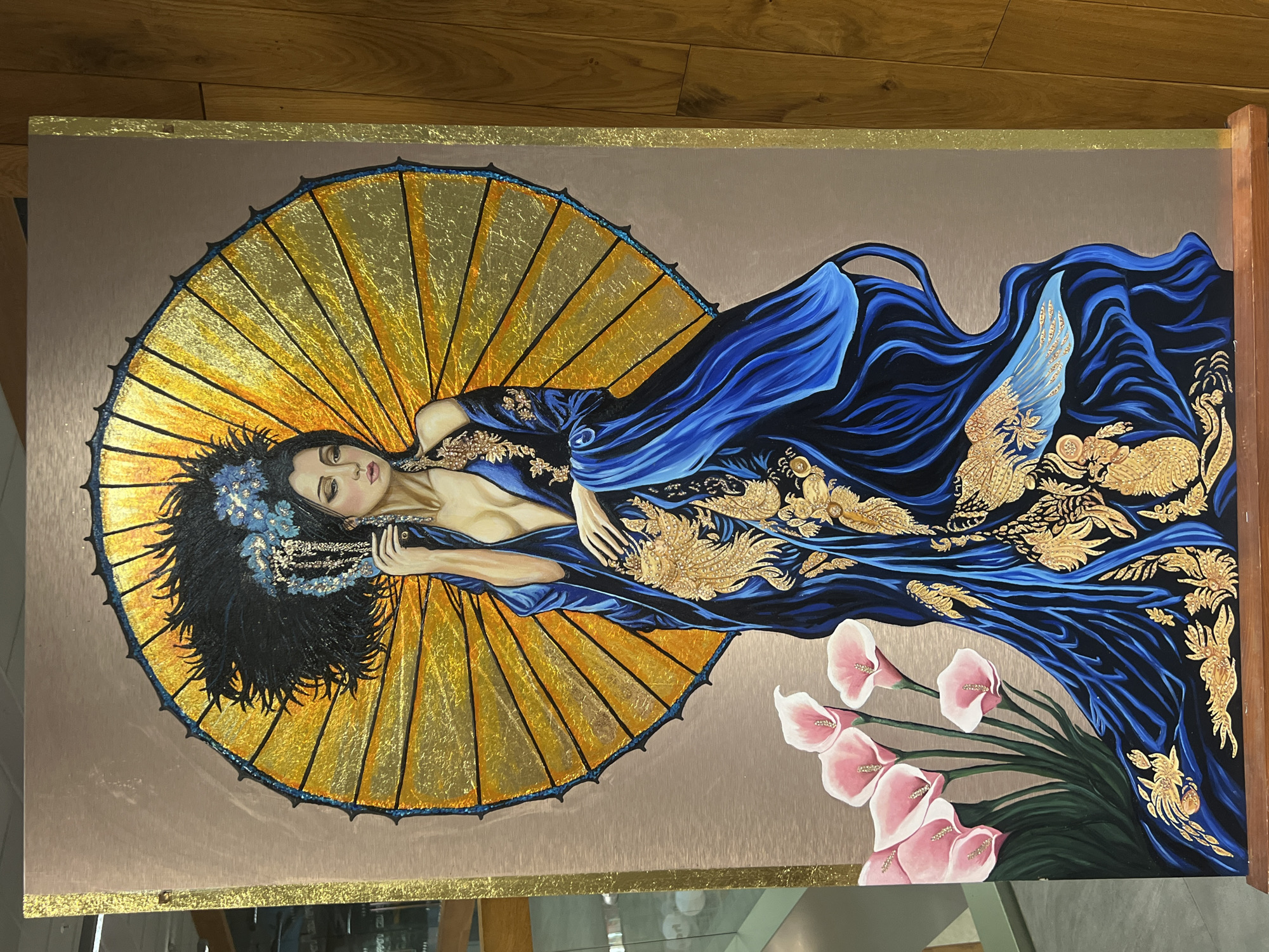 This media file is a painting of a woman reclining on a golden fan. The woman has dark hair adorned with blue flowers and is wearing a flowing blue robe decorated with gold embellishments. She appears to be asleep or in a meditative state, with her eyes closed and a peaceful expression on her face. The fan is large and circular, with golden spokes and a dark blue edge. Behind the woman, the robe drapes dramatically, creating a sense of movement and fluidity. To the right of the woman, a bouquet of pink calla lilies with green stems and leaves is depicted. The background of the painting is a neutral, textured color, possibly representing a wall or a fabric. The overall style of the painting is elegant and opulent, with a focus on rich colors and metallic accents. There is no visible text in the image.