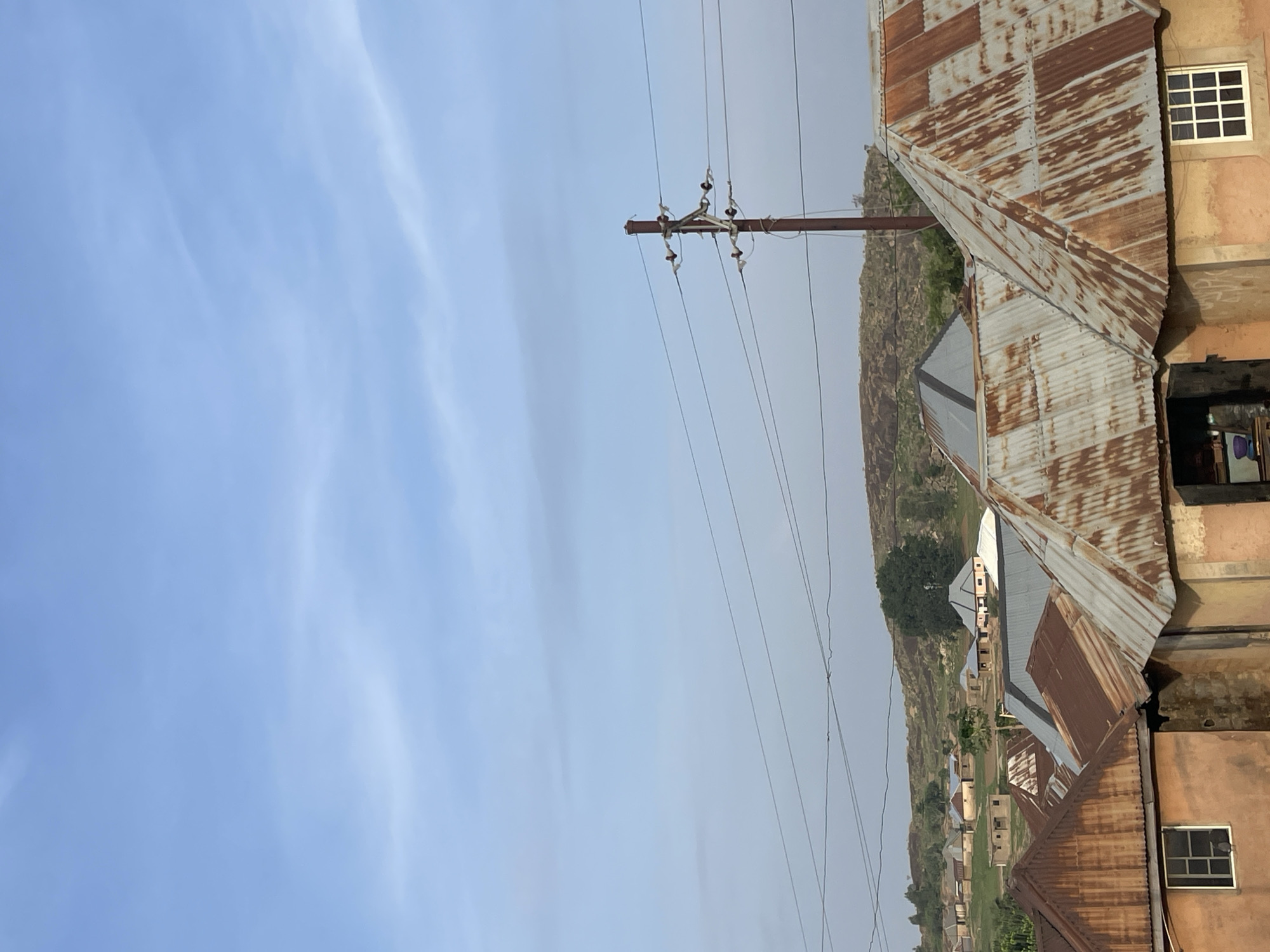 The image shows an outdoor scene in Barikin Gidar Kanat, Nigeria. The sky is mostly clear with light clouds. In the foreground, there are rooftops of buildings covered with corrugated metal sheets, many of which are rusted. A power pole with electrical lines is visible. In the distance, there is a grassy area with more buildings and a rock formation with sparse vegetation.