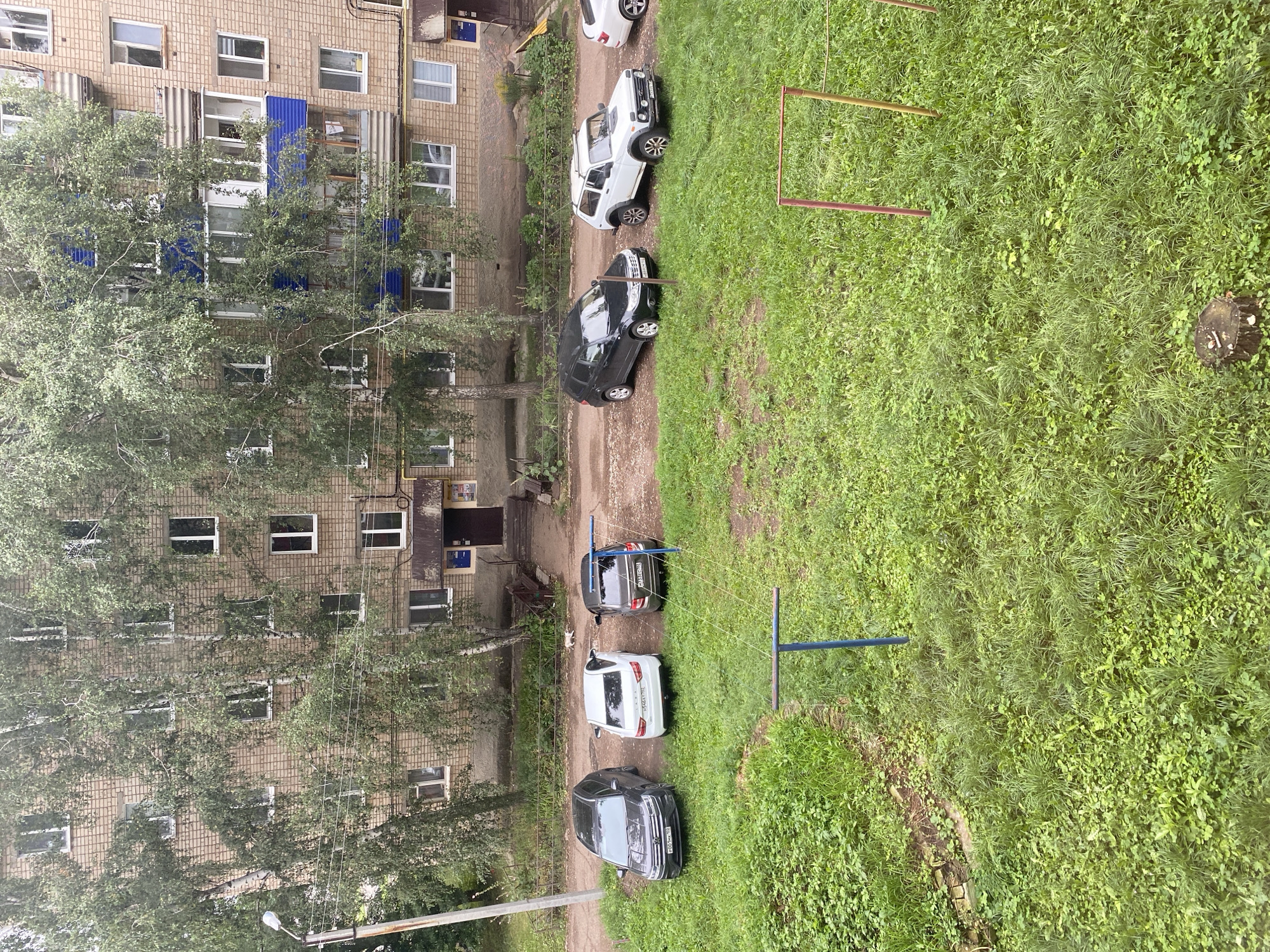 An exterior, high-angle, eye-level shot captures a parking area in front of a multi-story brick apartment building in Ishimbay, Russia. Trees with dense green foliage partially obscure the building's facade, which features numerous windows. Six cars are parked in a line on a dirt and gravel surface adjacent to a grassy area. From left to right, the cars are a black SUV, a white sedan, a dark gray sedan, a black SUV, a white off-road vehicle, and a partially visible white car. A blue clothesline pole stands in the grassy area, and a red and yellow structure, possibly part of a playground, is also visible. A small white dog walks in the distance near the building entrance. The environment appears to be overcast.