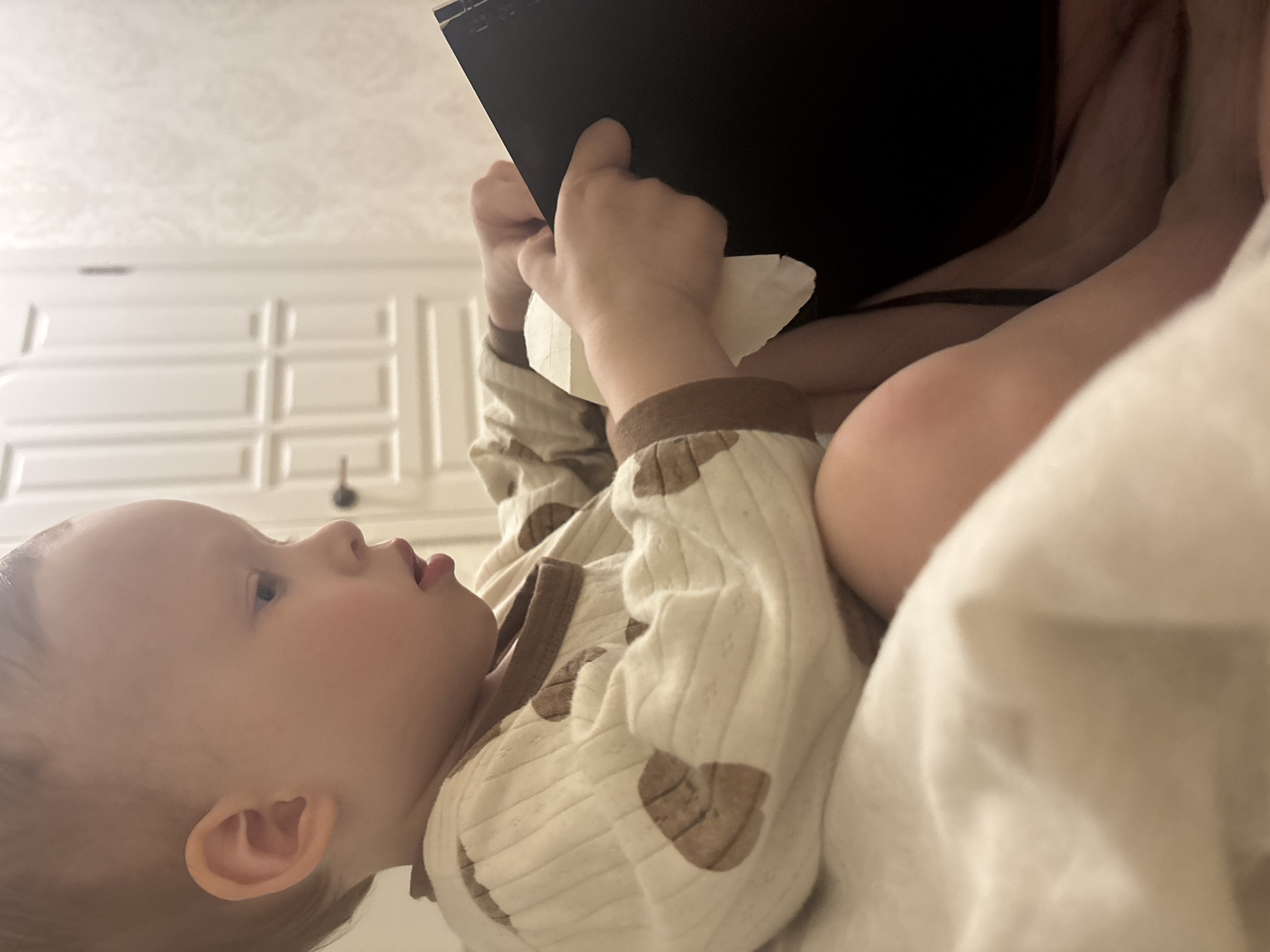 A young child, presumably in a residential setting in Ферма, Russia, is lying on a bed under a white blanket, facing left. The child is wearing a light-colored, patterned onesie with brown trim. The child's hands are holding a black object, possibly a book or tablet. In the background is a white paneled wall, potentially part of a cabinet or wardrobe.