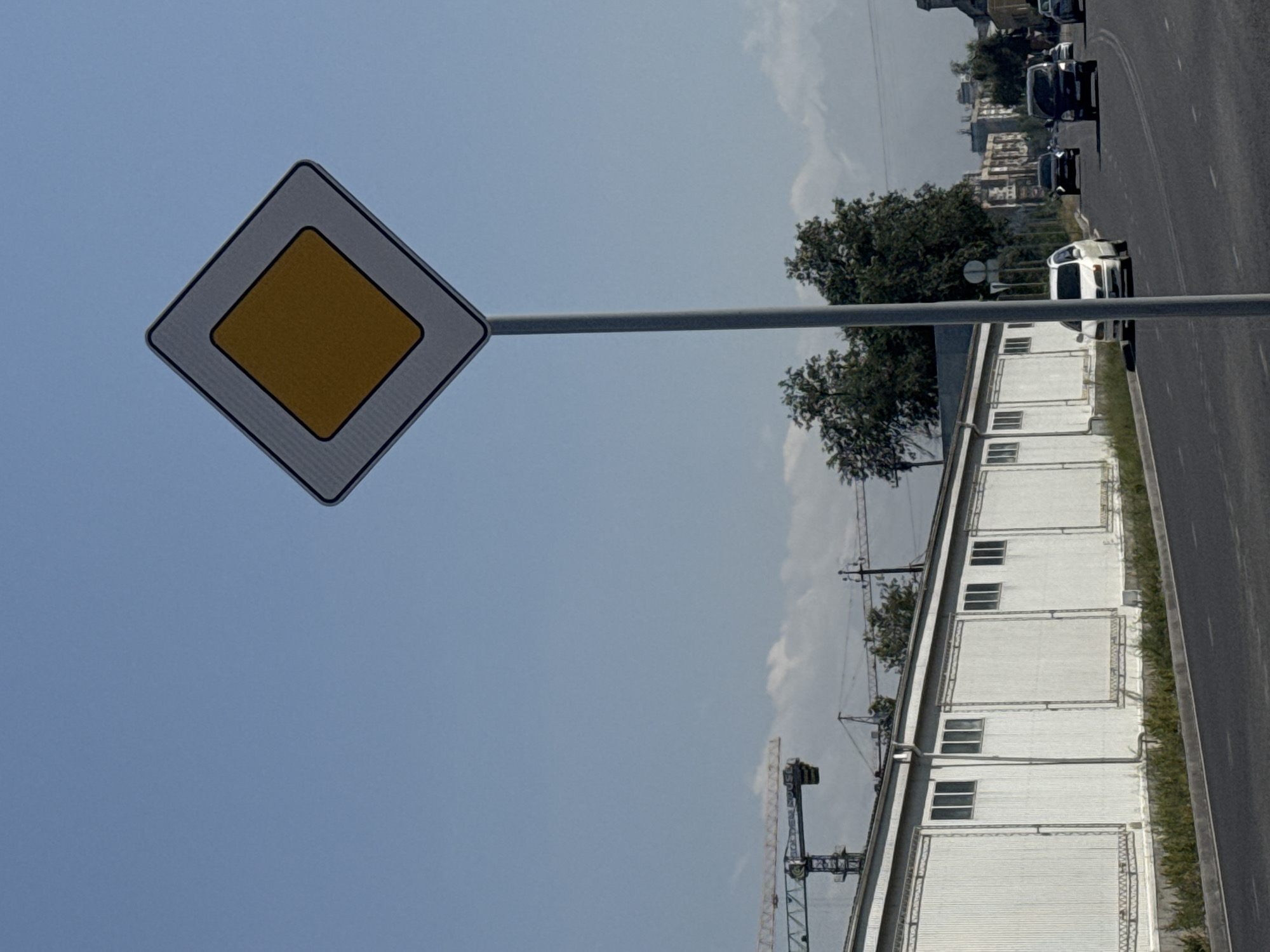 A street scene in Bishkek City, Kyrgyzstan, on a clear day. A prominent diamond-shaped "main road" traffic sign stands on a tall pole in the foreground. To the left, a long, white, corrugated metal building with multiple windows spans the width of the image. In the background, several construction cranes are visible, suggesting ongoing development. Beyond the building and cranes, a line of trees separates the construction area from a distant cityscape with apartment buildings. The road in front of the building shows a white car parked on the shoulder and several other vehicles driving on the asphalt. The sky is a bright, clear blue.