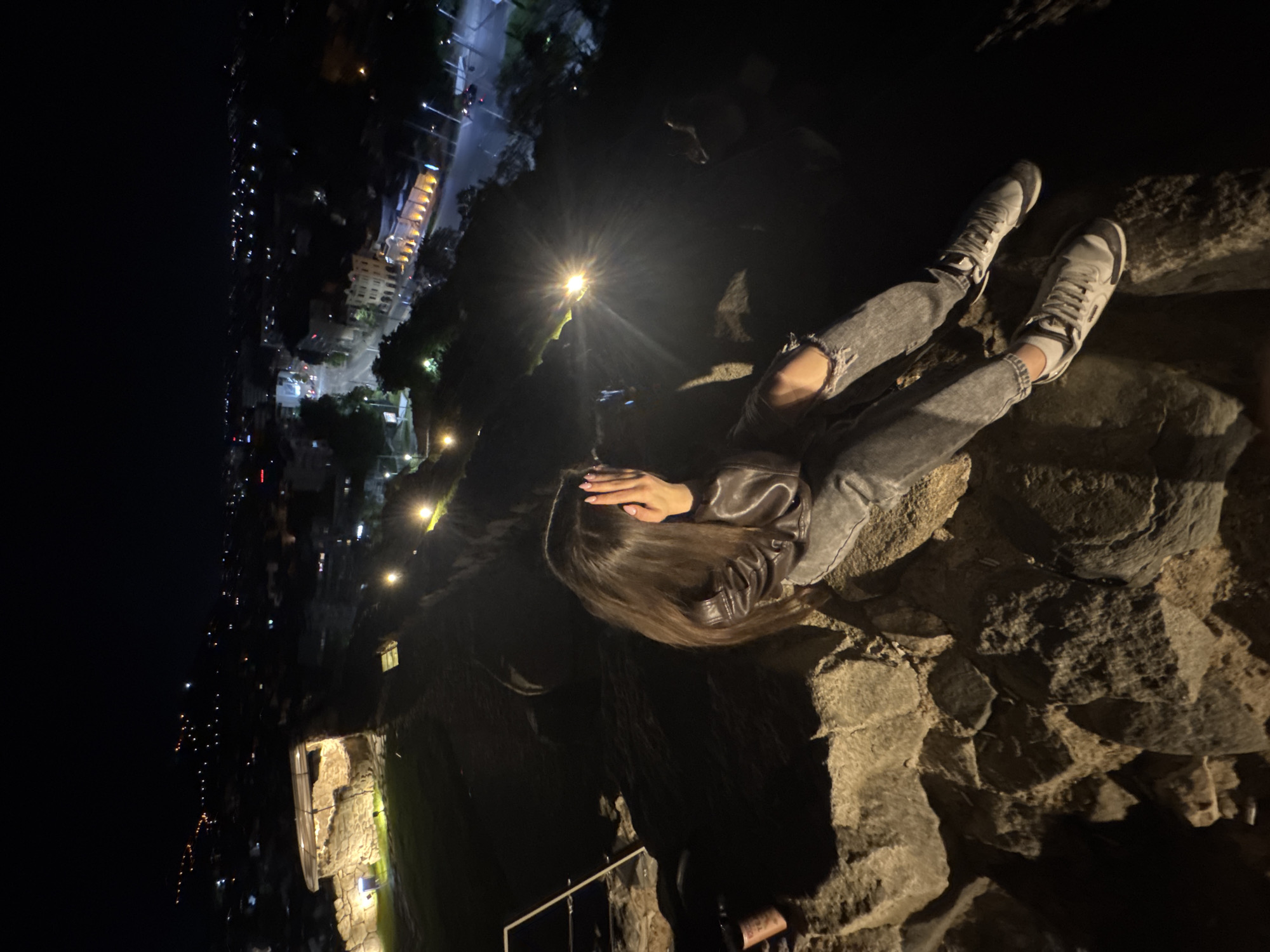 A vertically-oriented, low-light photograph taken at night depicts a woman sitting on a stone structure overlooking a city skyline.
The woman has long brown hair and is wearing a gray leather jacket and ripped jeans. She is positioned diagonally in the frame, with her back angled towards the viewer and her head tilted downward, her face obscured by her hand. Her legs are stretched out, resting on a stone surface. She is wearing white socks and gray athletic shoes.
The surrounding environment consists of a stone structure, with visible rocks and crevices. In the background, a city skyline is illuminated with artificial lighting, including street lamps and buildings. The overall ambiance is dark, but with concentrated light sources.
Based on visual cues, the location is potentially Nebet Tepe in Plovdiv, Bulgaria, a historic landmark with stone structures overlooking the city. There is a bottle next to the woman.