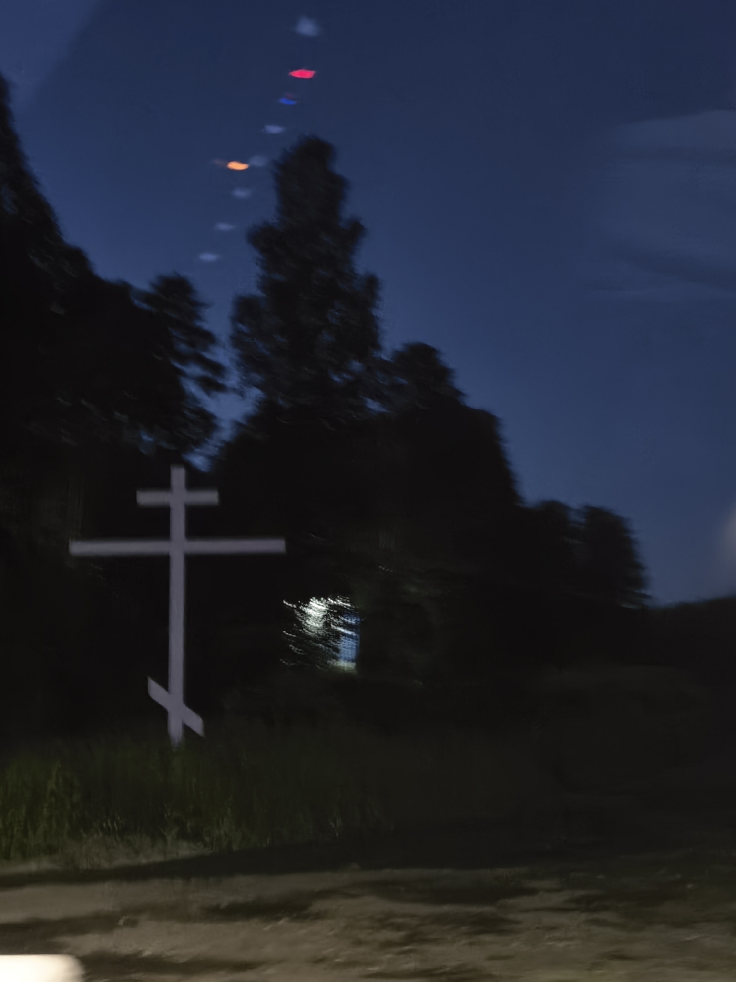 A blurry, dark photograph taken at night shows a white, eight-pointed Eastern Orthodox cross standing in a field of grass. The cross is positioned in the center-left of the frame. Behind the cross, silhouetted trees are visible against a dark blue sky. In the upper portion of the sky, several out-of-focus, colored lights are present, appearing as red, blue, and orange streaks. The overall impression is of motion blur, suggesting the image was taken from a moving vehicle. The scene is devoid of people.