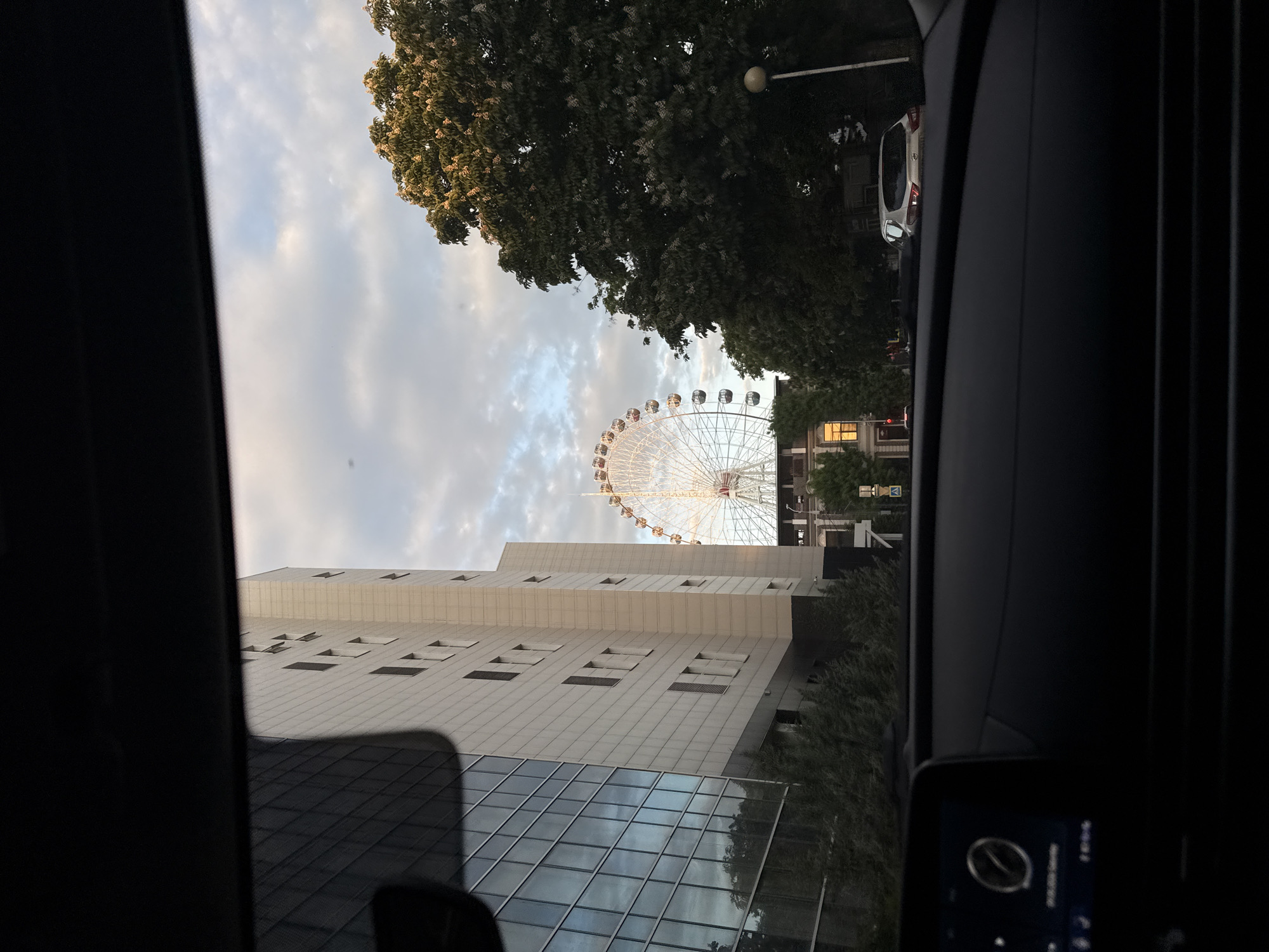 Captured from within a vehicle, the image presents a cityscape featuring a large Ferris wheel. A multi-story building with reflective glass and stone exterior stands prominently. Foliage fills the frame above the building, with overcast skies above. The Ferris wheel is visible between the building and trees. The scene is viewed through the car's windshield, with parts of the vehicle's interior visible on either side of the frame. A small screen with digital elements is visible on the right.