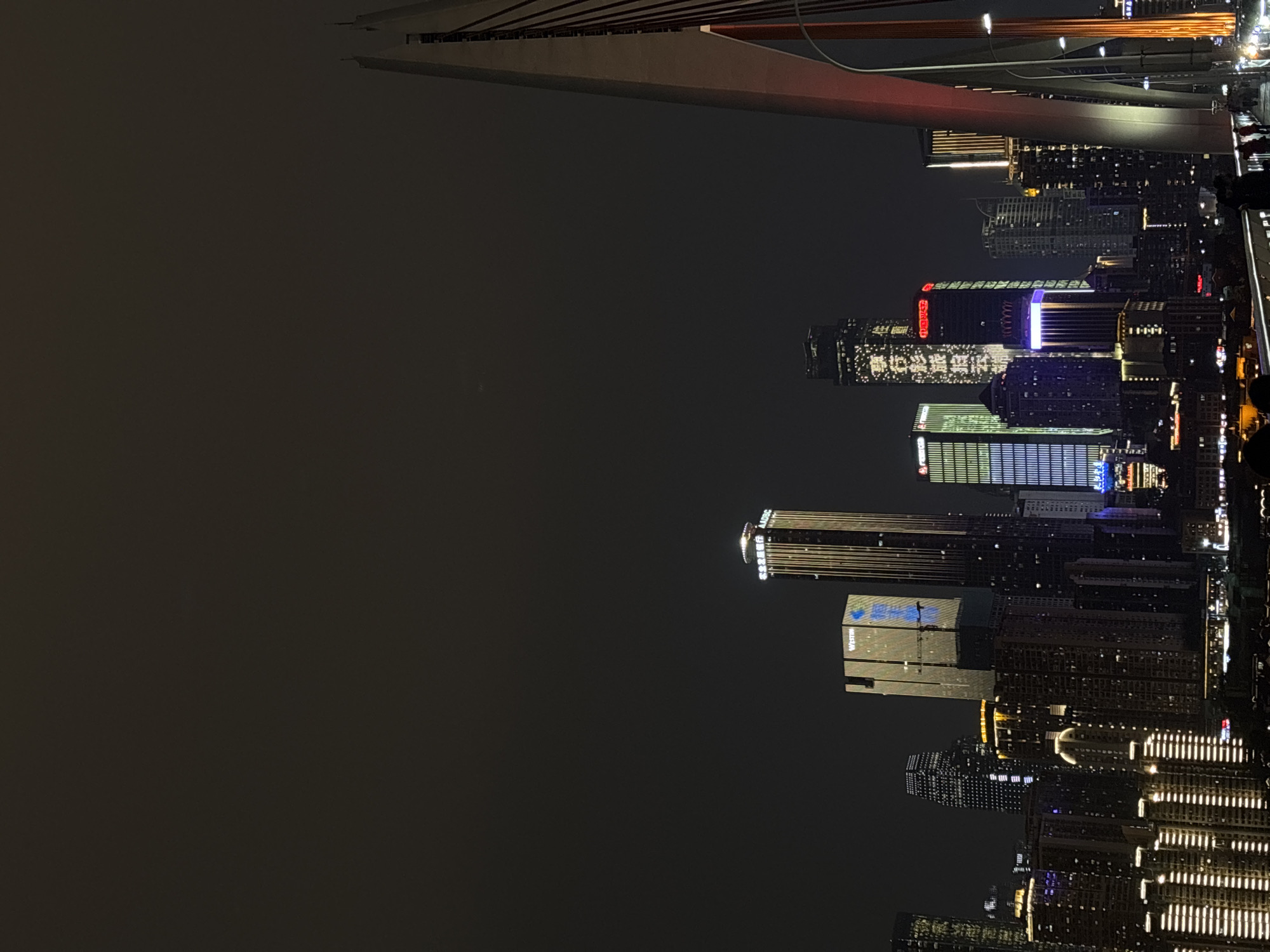 This media file captures a vibrant and densely illuminated cityscape at night. The scene is dominated by numerous high-rise buildings and skyscrapers, stretching across the frame under a dark sky.
In the upper part of the image, a large, modern bridge structure spans across the urban landscape. This bridge, possibly a cable-stayed or arch design, is illuminated with warm, reddish-orange lights along its supports and possibly its deck. On what appears to be a walkway or viewing platform on the bridge's right side, a few small, indistinct figures are visible, suggesting people are present to observe the night view or traverse the bridge.
The city below the bridge is a tapestry of light, with countless windows glowing and building facades adorned with decorative lighting. Various architectural styles are visible among the high-rises. Several buildings feature prominent illuminated signage. Most notably, a tall skyscraper near the center prominently displays the Chinese characters "重庆来福士" (Chongqing Raffles City) and, below it, the message "重庆欢迎你" (Chongqing welcomes you). Further to the right, another building exhibits the "WESTIN" logo in English, along with the Chinese characters "威斯汀" (Weisiting), indicating a Westin hotel. Other buildings display animated light patterns and corporate branding, contributing to the city's dynamic nocturnal appearance.
The time of day is clearly night, with no natural light visible. The atmosphere is one of bustling urban energy and impressive modern development. The explicit text on the buildings confirms the location as Chongqing, China. Given the context of Nan'an District, the view is likely from a vantage point overlooking one of Chongqing's rivers, with the lit bridge and the high-rises creating a spectacular evening panorama.
