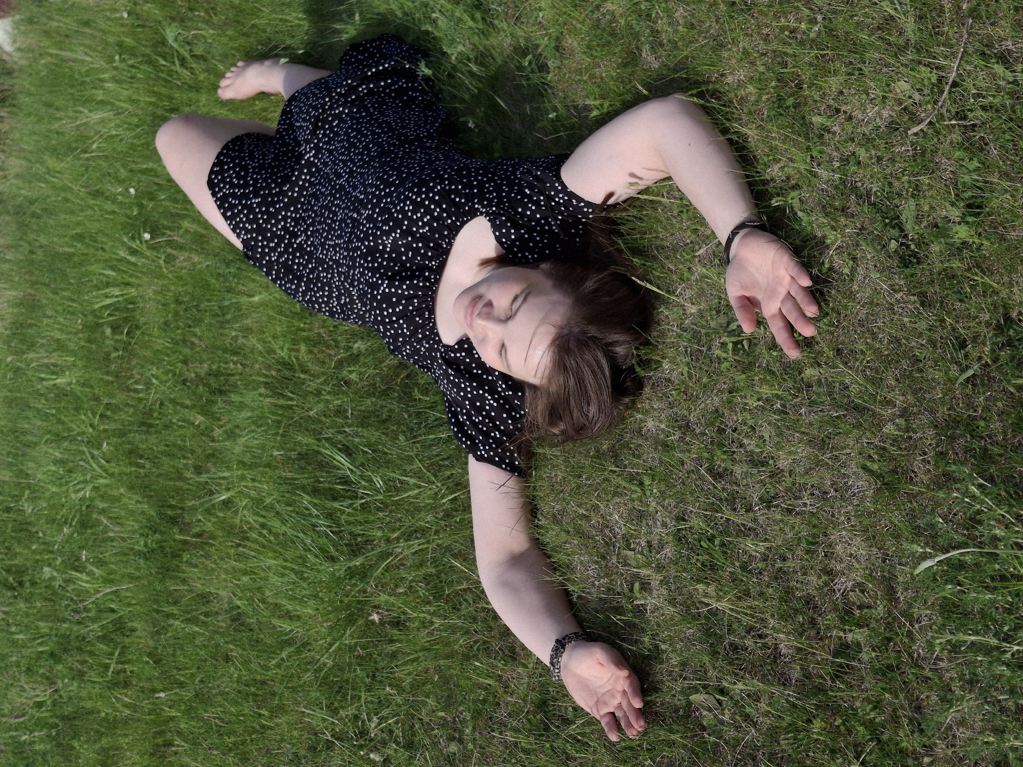 A young woman lies on her back in a grassy field, arms outstretched. She wears a short, black dress with white polka dots, and bracelets on both wrists. Her eyes are closed. The location is most likely a grassy field in or around the city of Ольховка, Russia.